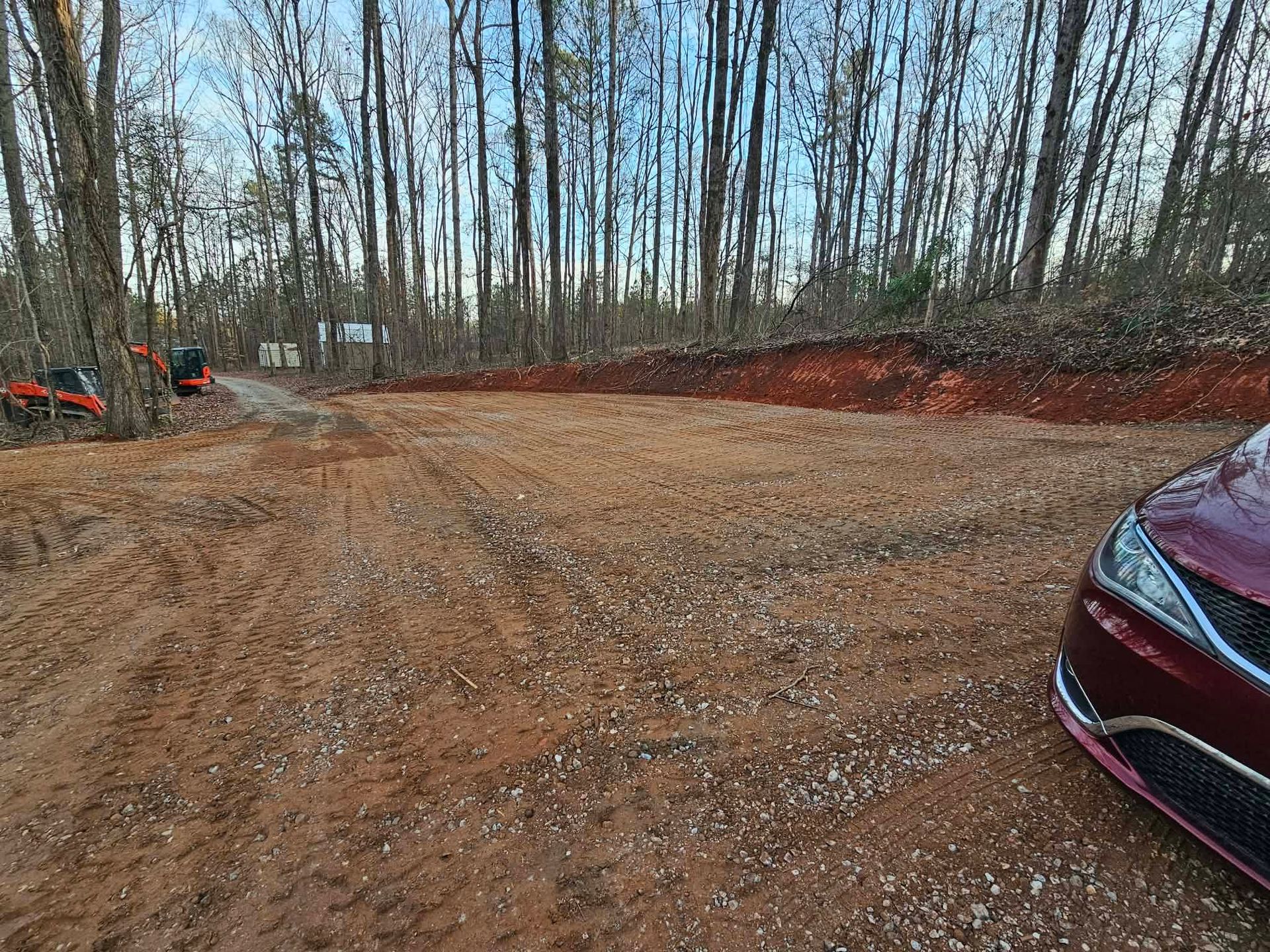 Gravel parking area by leafless trees, with a maroon car at right and orange equipment at left