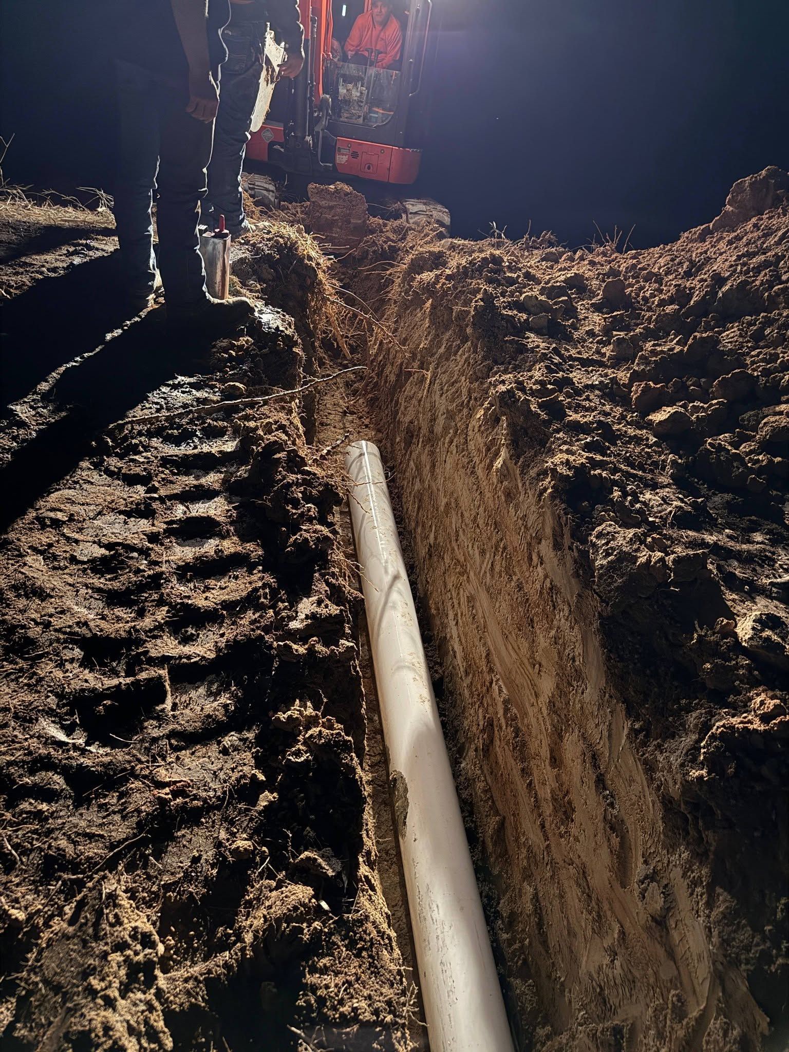 Worker standing in a trench with a long white pipe laid in the soil at night