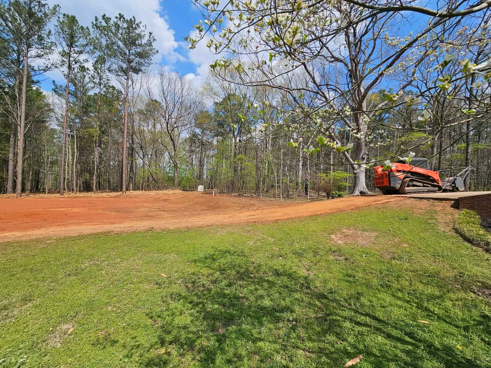 Orange excavator grading red dirt beside a grassy field and trees under a blue sky