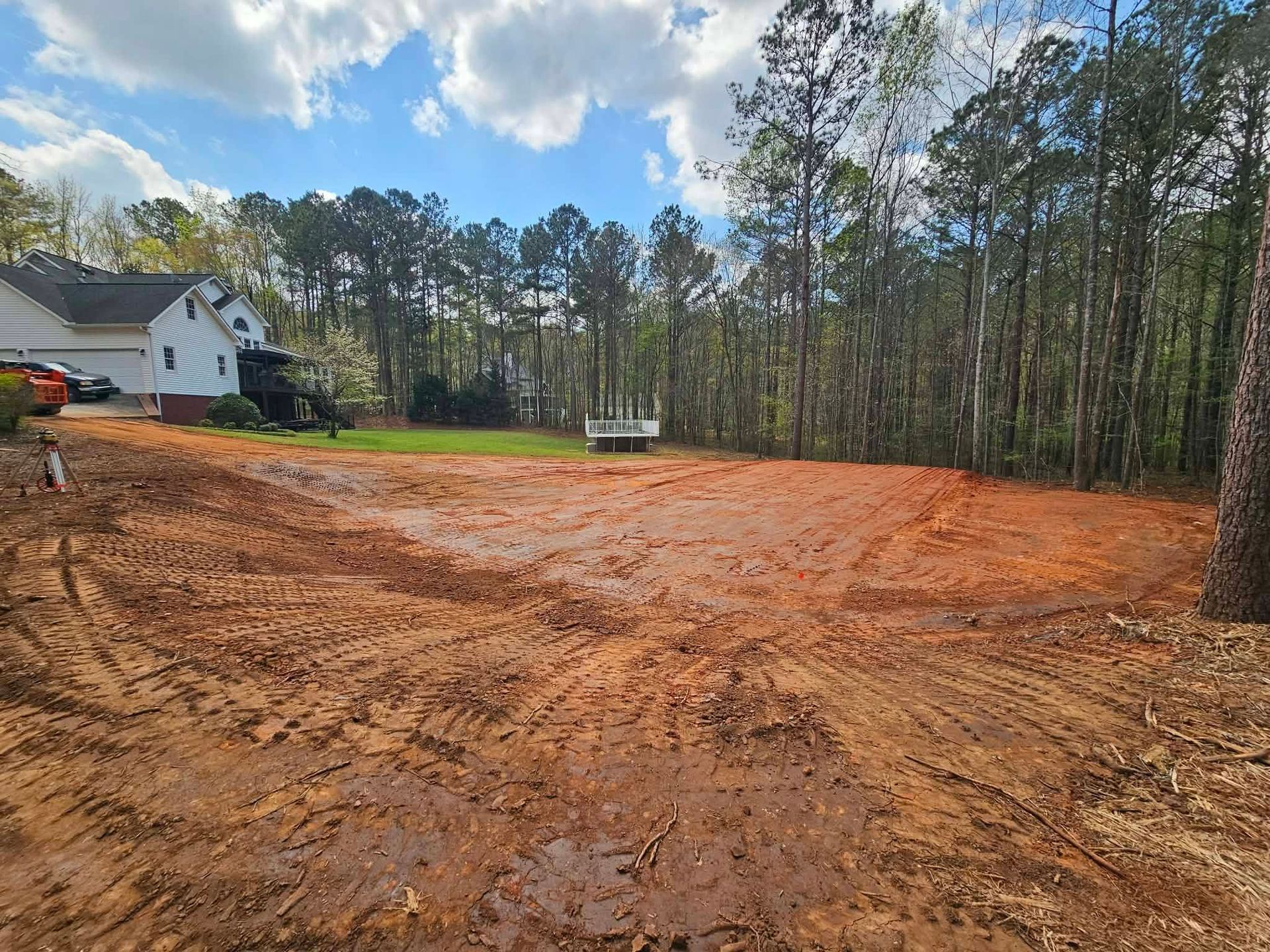 Cleared dirt lot beside a house and trees under a partly cloudy sky