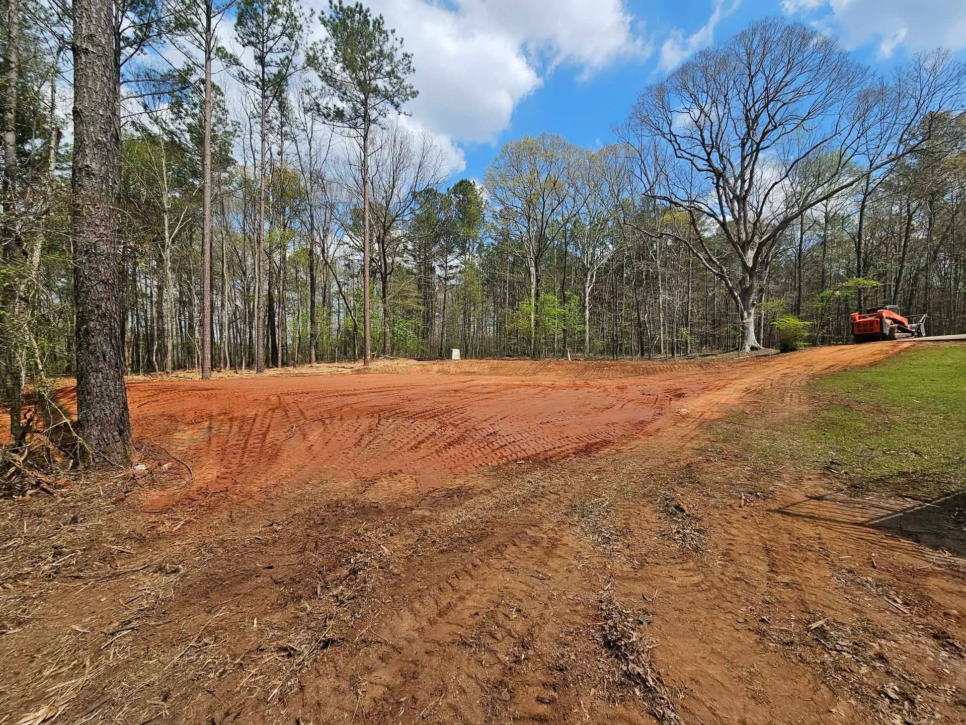 Dirt clearing in a wooded area with a small orange excavator in the distance