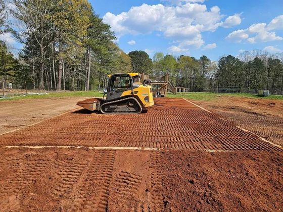 Yellow skid steer grading a red dirt lot with trees and a blue sky in the background