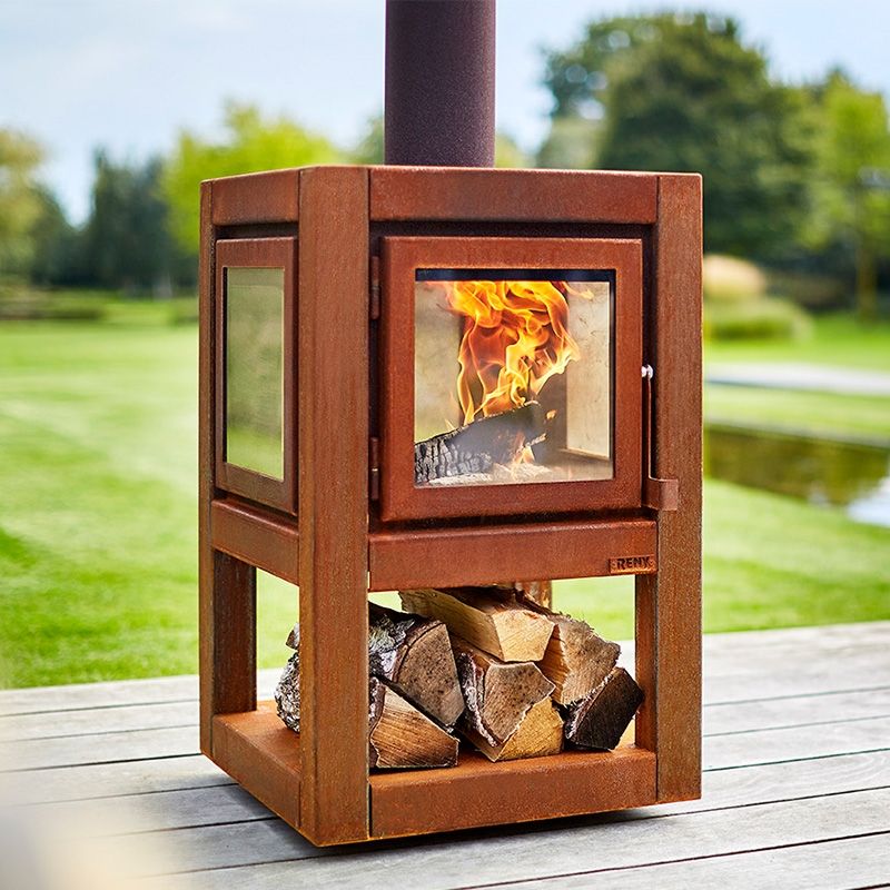 Rust-colored outdoor wood stove with a visible fire and stacked logs beneath, on a patio by a grassy lawn