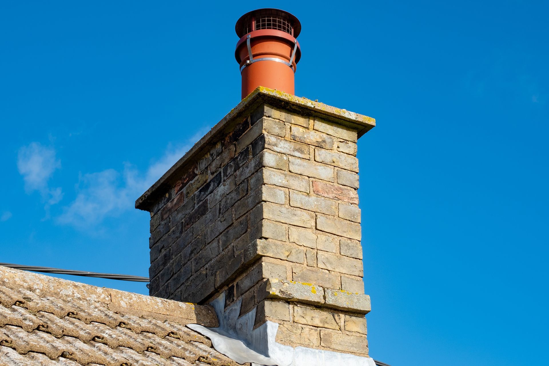 Brick chimney with red metal cap on stone house roof under clear blue sky, residential home exterior.