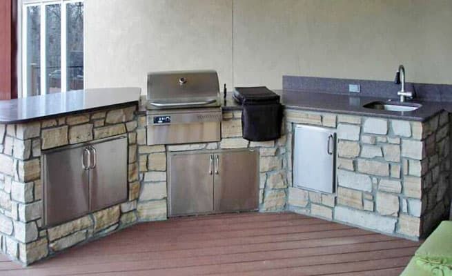 Outdoor stone kitchen with stainless grill, sink, and cabinets on a wooden deck