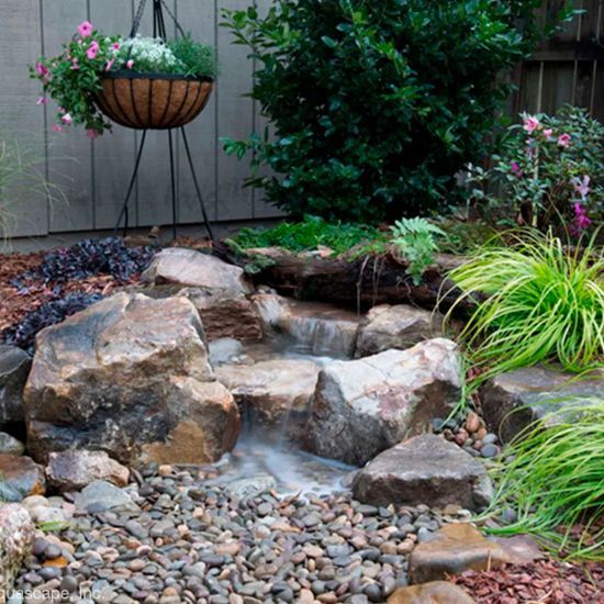 Small garden waterfall with rocks, pebbles, and lush green plants beside a wooden fence