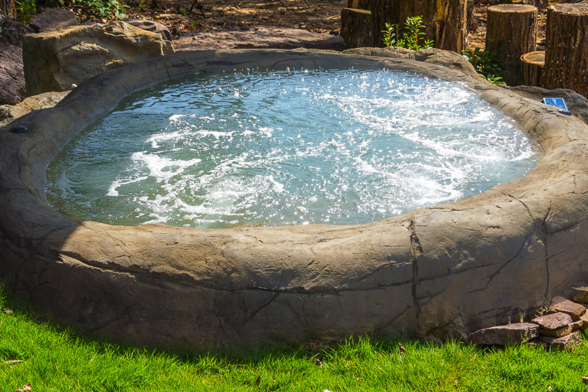 Round stone hot tub filled with bubbling blue water outdoors on a grassy patio