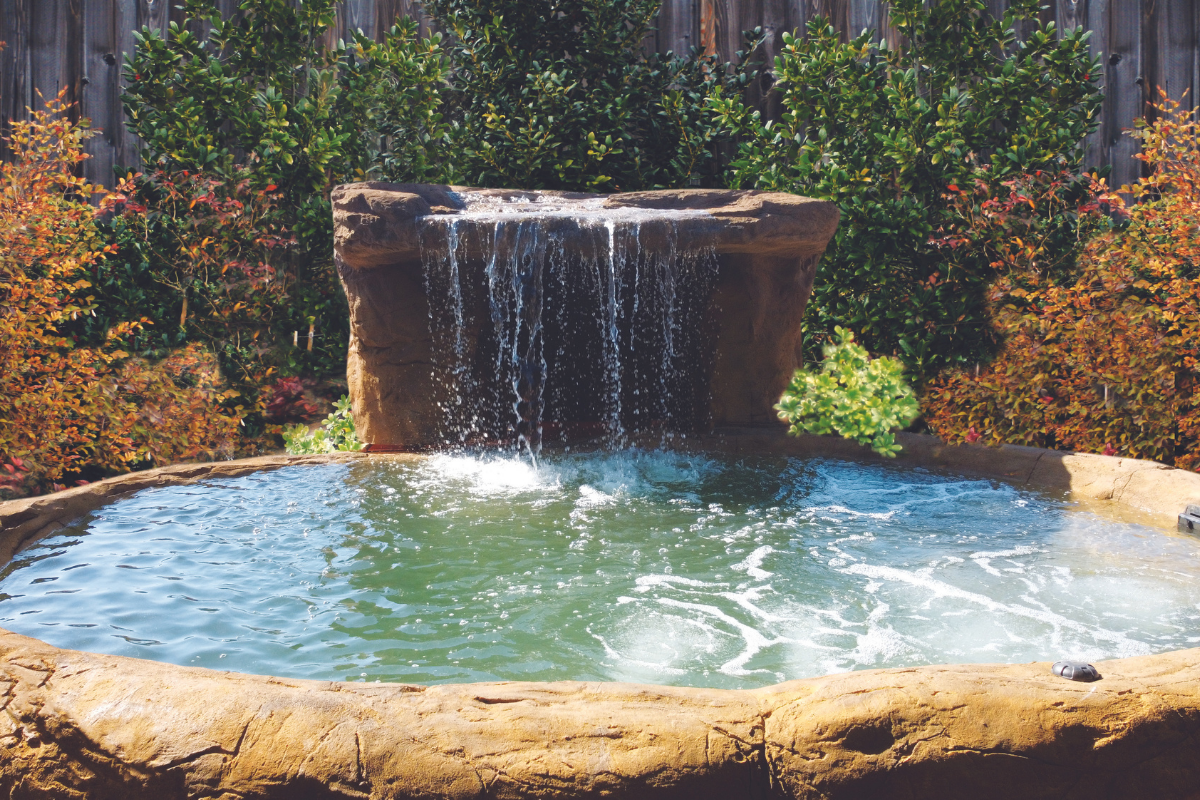 Small waterfall spilling into a round blue pool surrounded by stone and autumn foliage