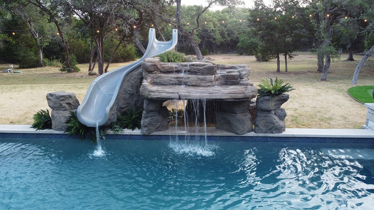 Backyard pool with a rock waterfall and a gray slide surrounded by trees