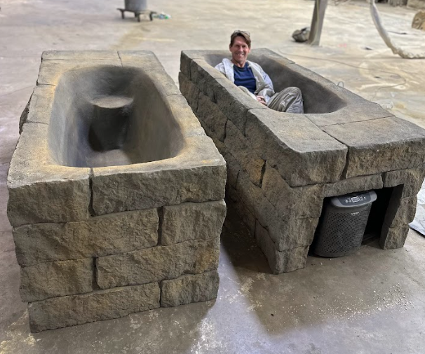 Person sitting inside a stone bench shaped like a coffin on a paved outdoor area.