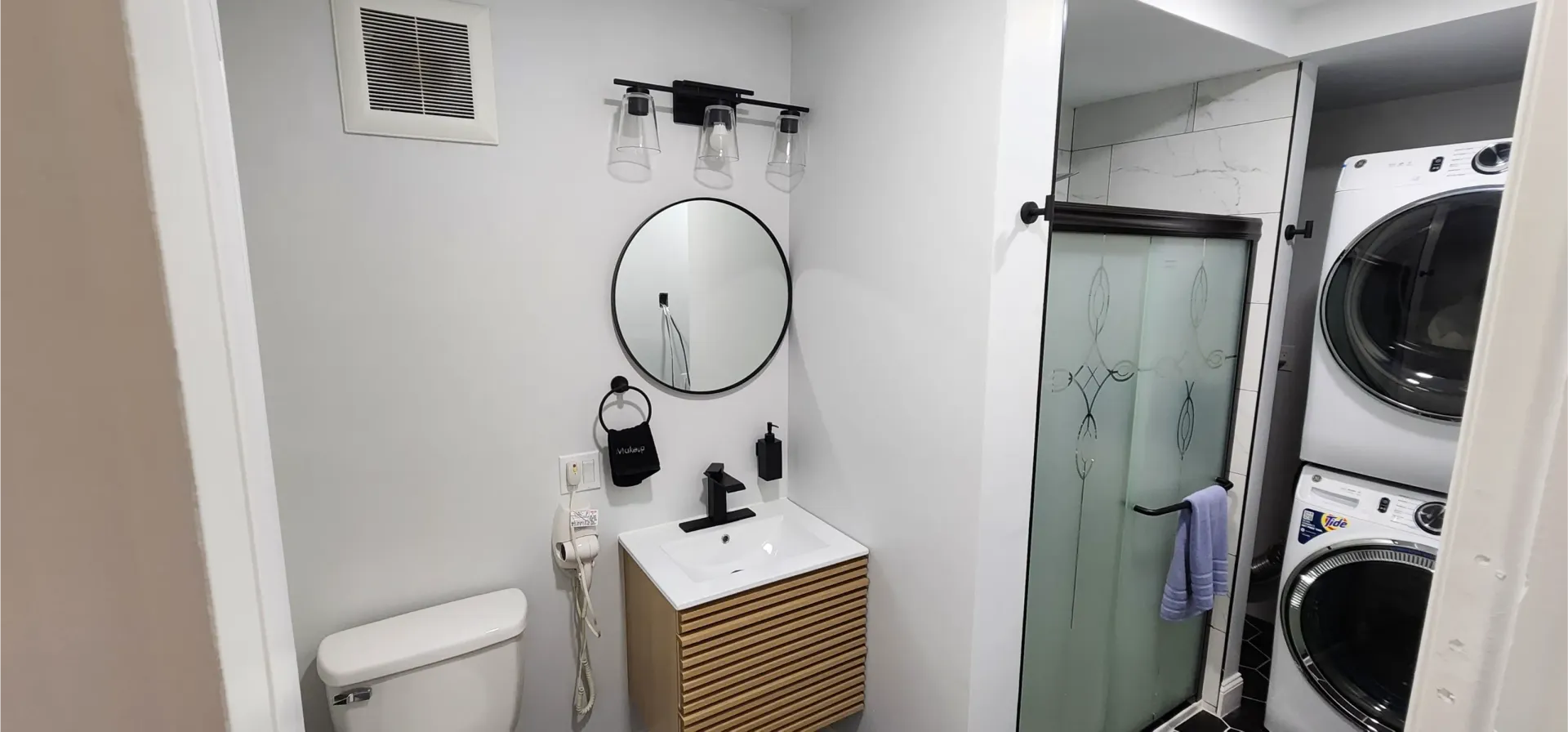 A bathroom with a white toilet, modern vanity, round mirror, and a stacked washer and dryer unit next to a shower.