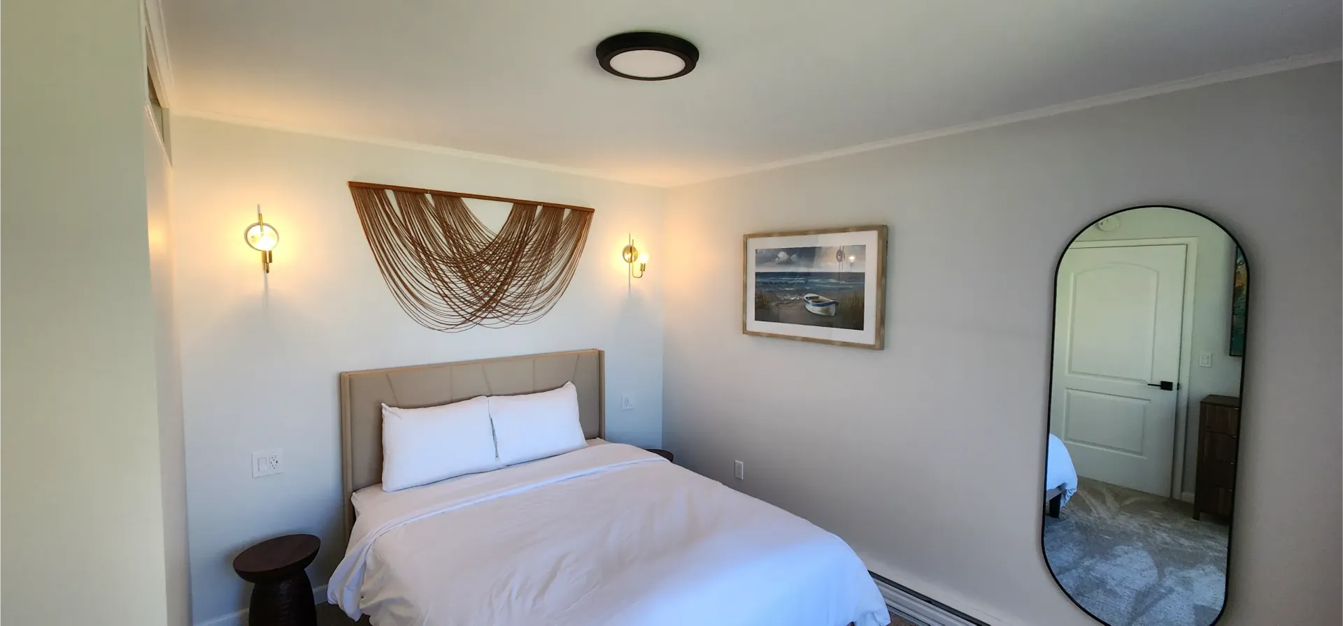 A neutral-toned bedroom with a bed, macramé wall art, two small wall sconces, a framed picture, and an arched floor mirror.