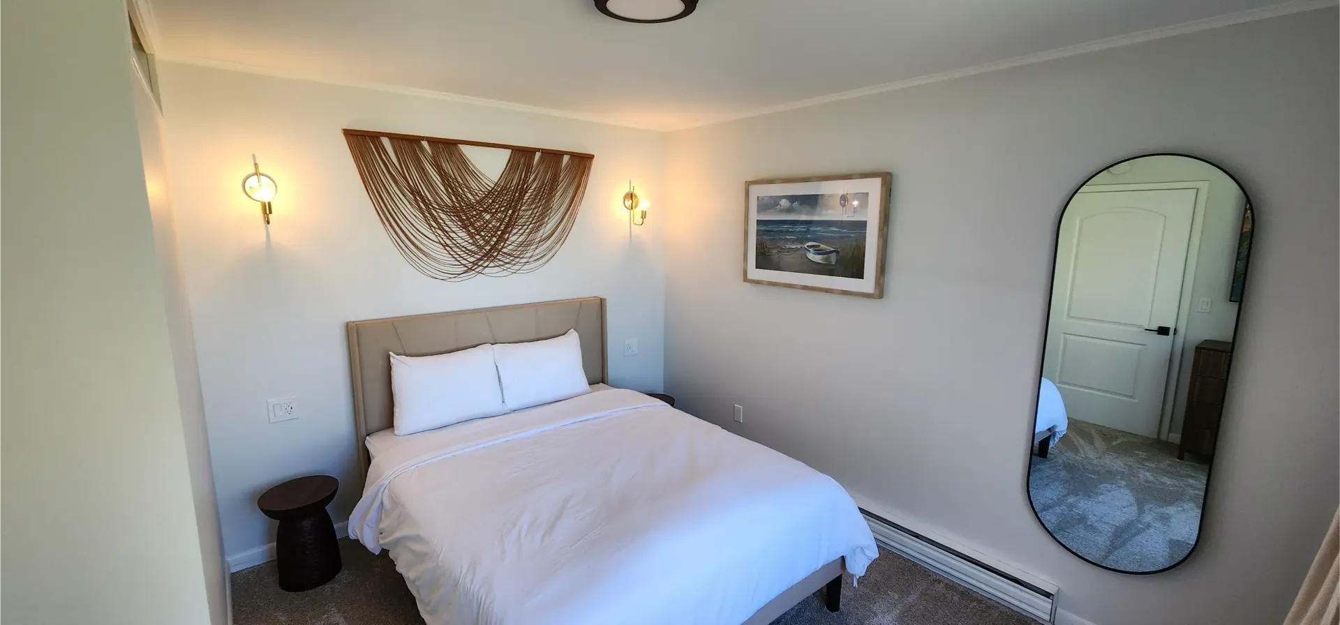 A bedroom featuring a bed with white bedding, a decorative wall hanging, wall sconces, a framed landscape, and a mirror.