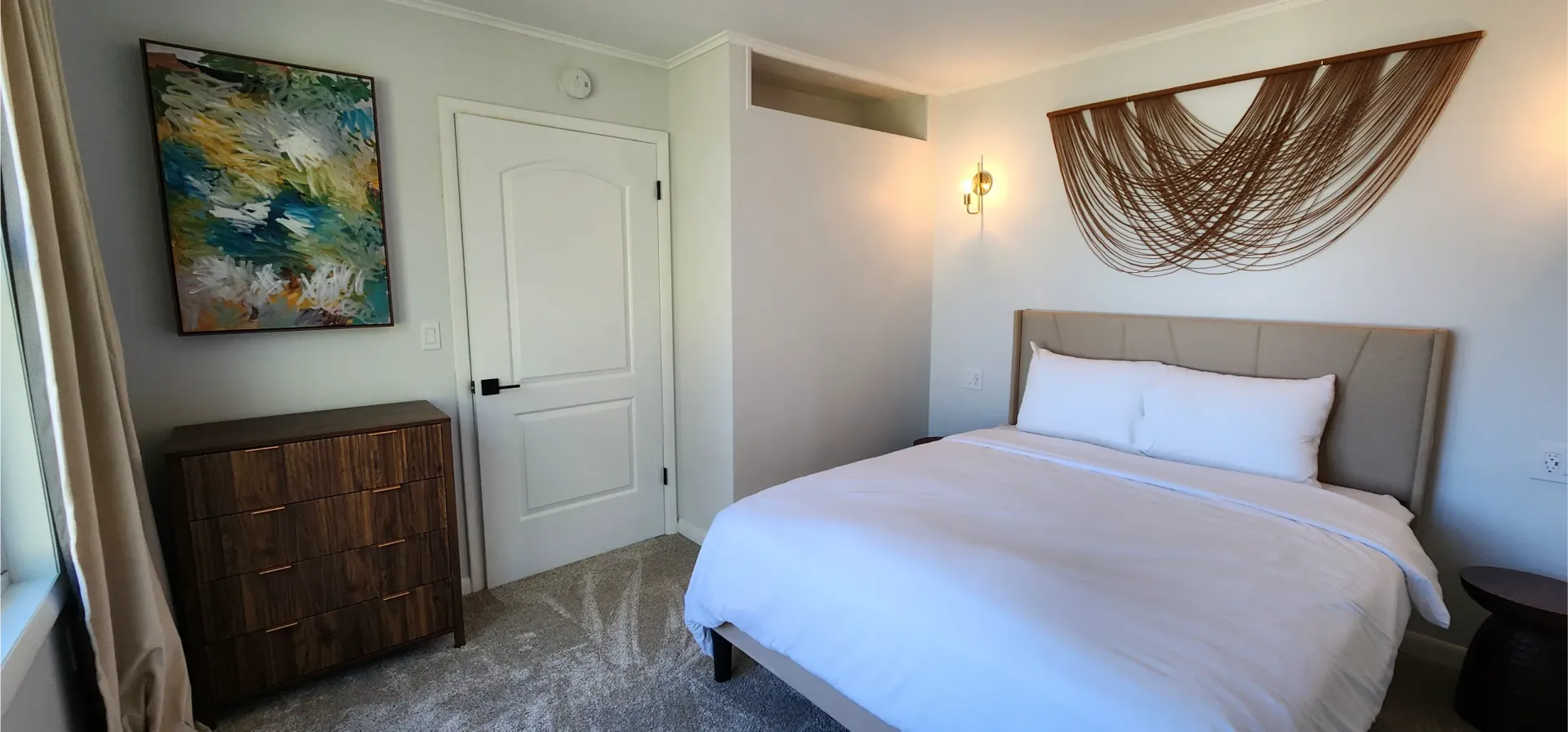 A bedroom with a queen bed, white linens, wood dresser, wall art, and a decorative macramé piece above the headboard.