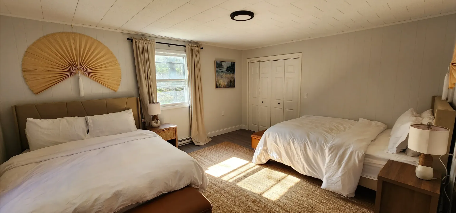 A bedroom with two beds, white bedding, a wooden fan wall hanging, and a window with curtains.