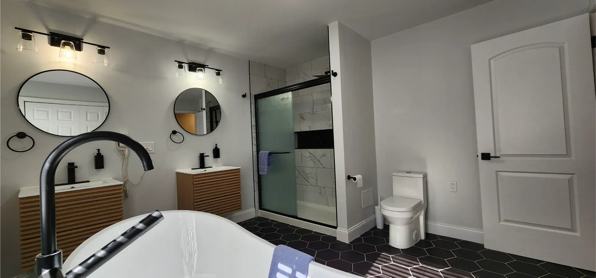 A modern bathroom with a freestanding white tub, two circular mirrors over wood-patterned vanities, and a glass shower.