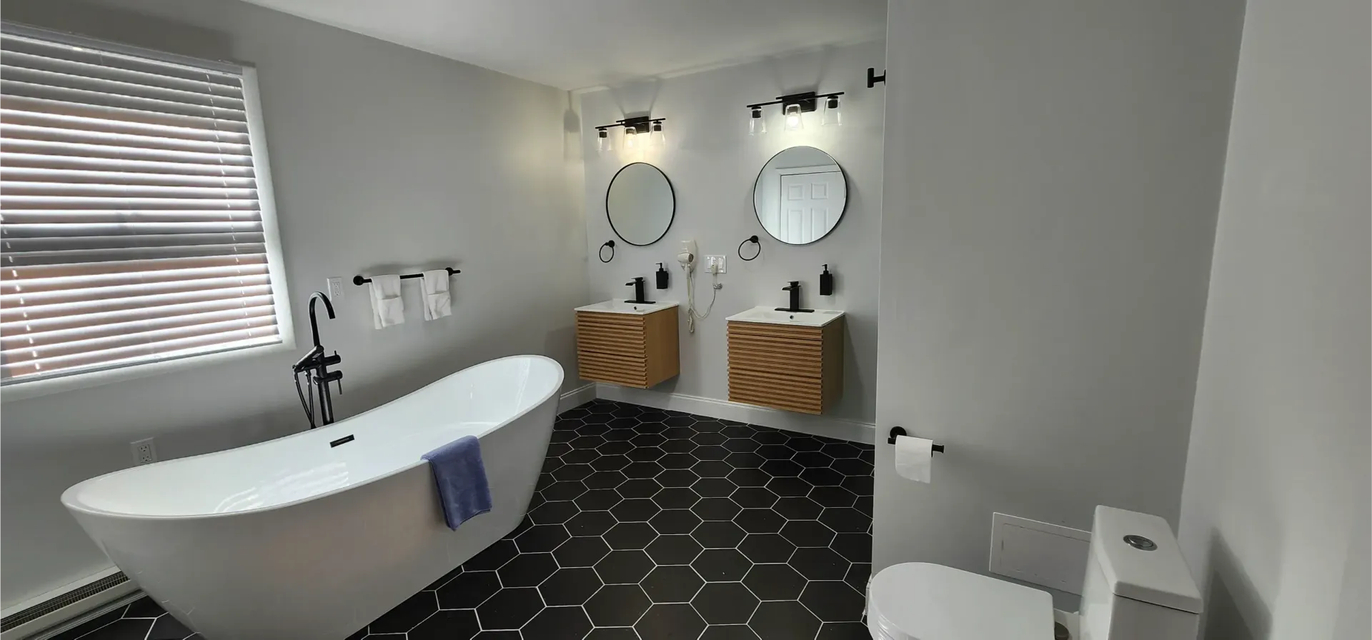 Modern bathroom with a freestanding tub, dual wooden vanities with round mirrors, and black hexagonal floor tiles.