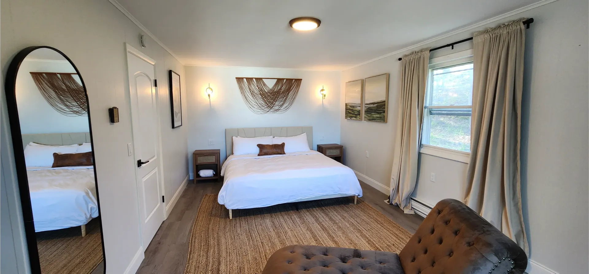 A bright bedroom with a king bed, natural wood accents, a round mirror, and a tufted chair on a woven area rug.