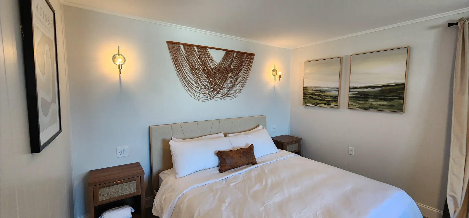A modern bedroom featuring a bed with white bedding, wooden nightstands, wall-mounted sconce lights, and wall art.