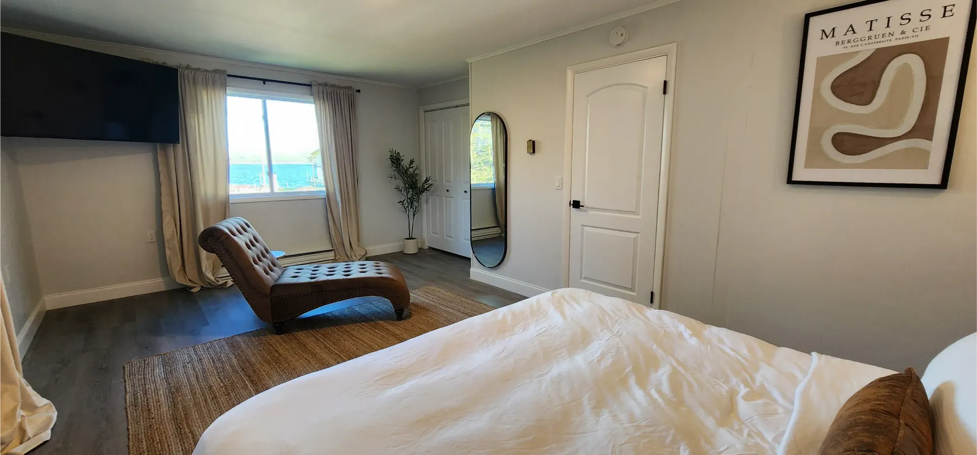 A modern, bright bedroom features a white bed, a chaise lounge, a wall-mounted TV, a floor mirror, and framed art.