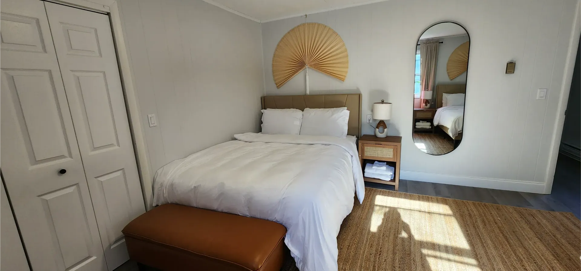 A bedroom featuring a bed with white bedding, a leather bench, a woven fan wall decoration, and an oval mirror.