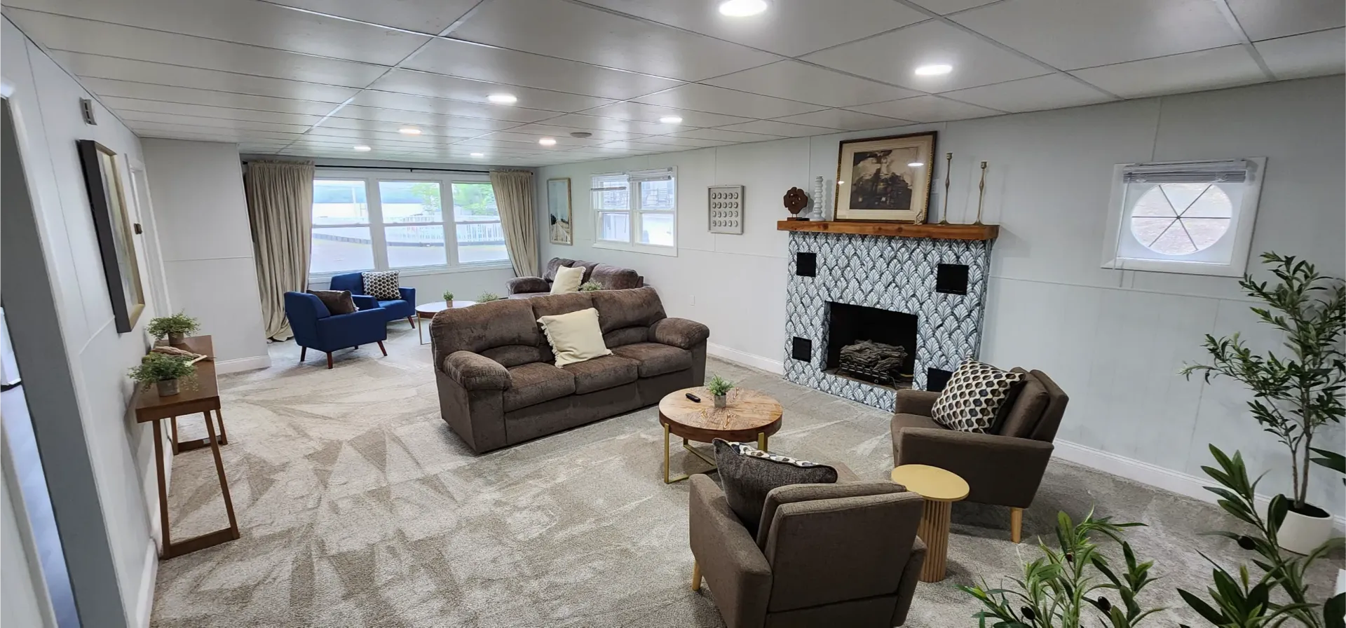 A spacious, light-toned living room featuring a fireplace, a brown sofa, two brown armchairs, and a drop-ceiling.