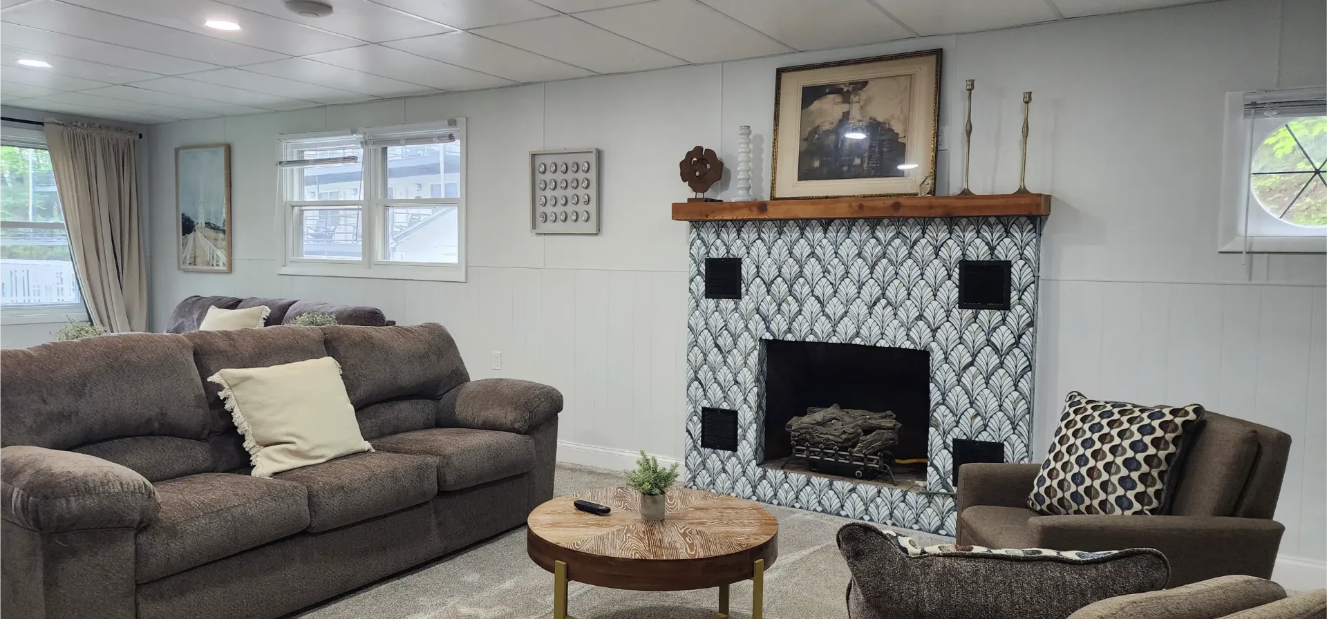A cozy living room with a patterned fireplace, a brown sofa, an armchair, and a circular coffee table on a light rug.