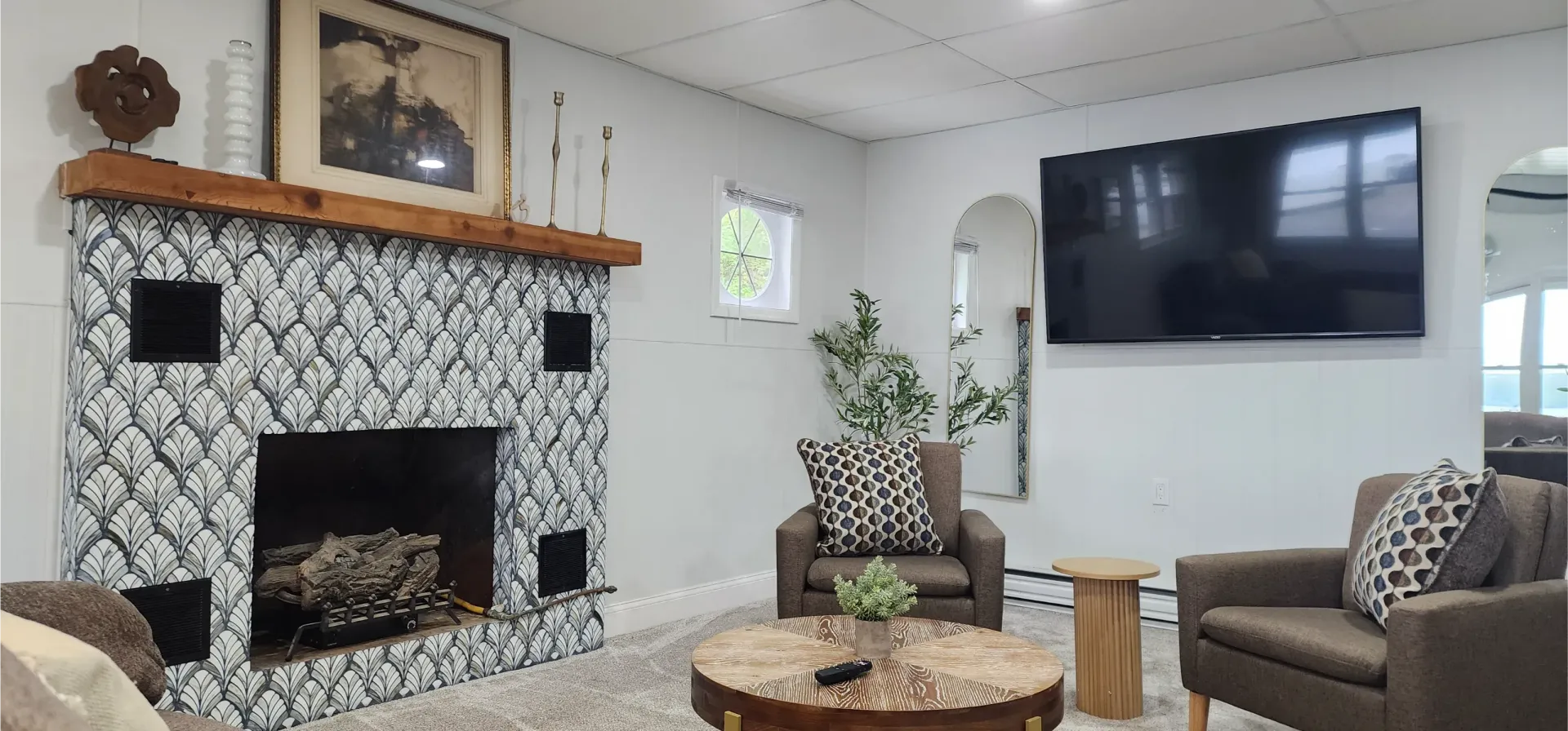 A living room featuring a patterned tile fireplace with a wooden mantel, two armchairs, a round table, and a wall-mounted TV.