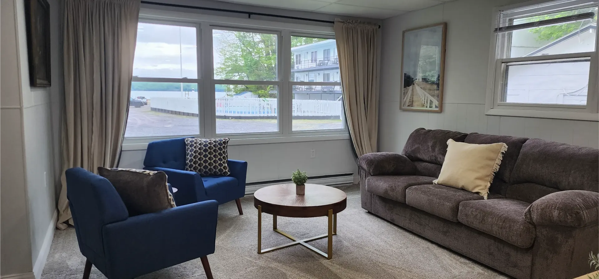 A living room with a brown sofa, two blue armchairs, a round coffee table, a patterned rug, and curtains by a large window.