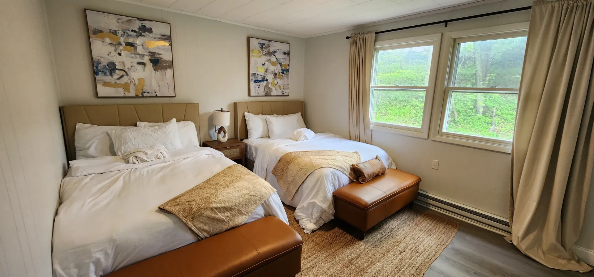 A bedroom features two queen beds with white bedding, gold throw blankets, brown benches, and artwork on a light wall.