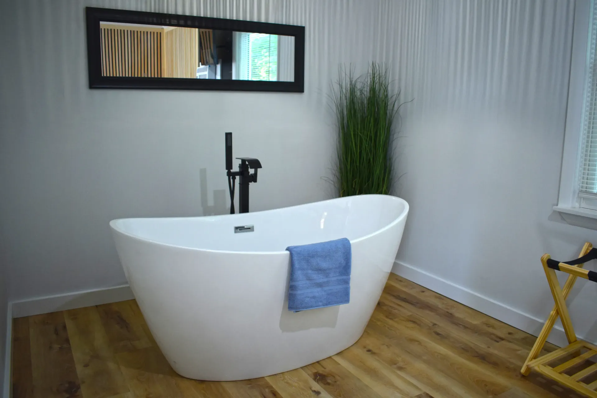 A white freestanding bathtub with a black faucet and a blue towel sits on a wood floor in a room with light walls.