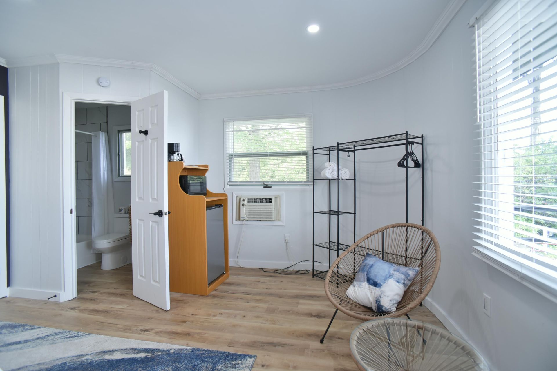 A brightly lit room with light wood floors, a small bathroom, a wooden cabinet, metal shelving, and wicker chairs.