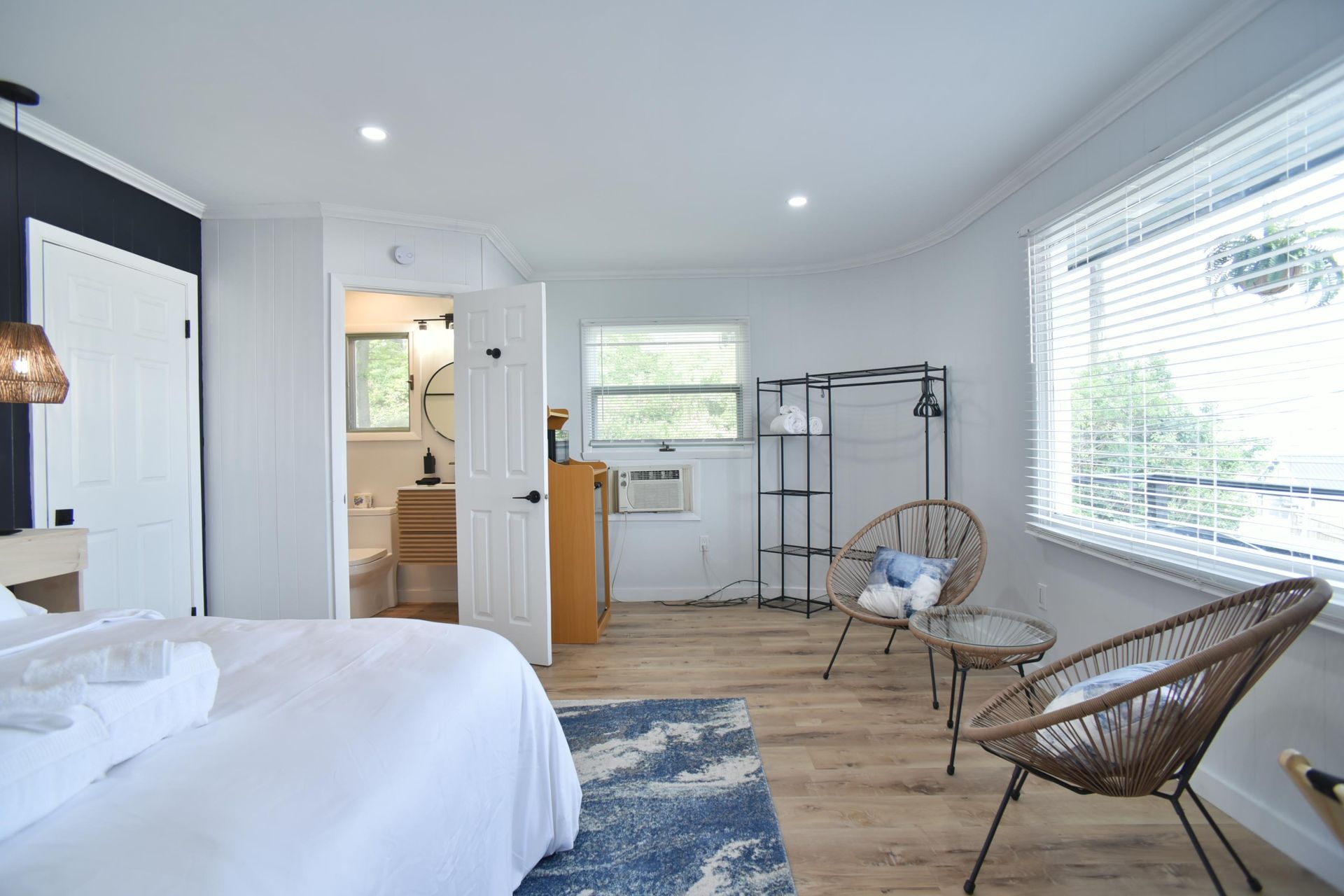 A modern bedroom with a large bed, two wicker chairs, a small table, a metal rack, and an open door to a bathroom.