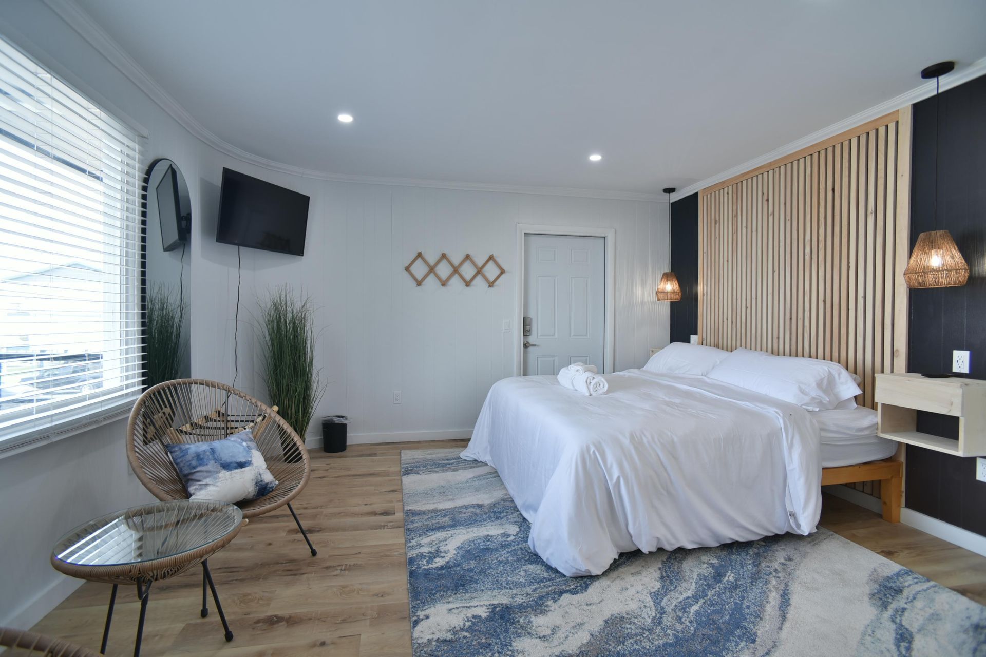 Modern bedroom with a white-bedded queen size bed, wood panel headboard, rattan chair, glass table, and a blue area rug.