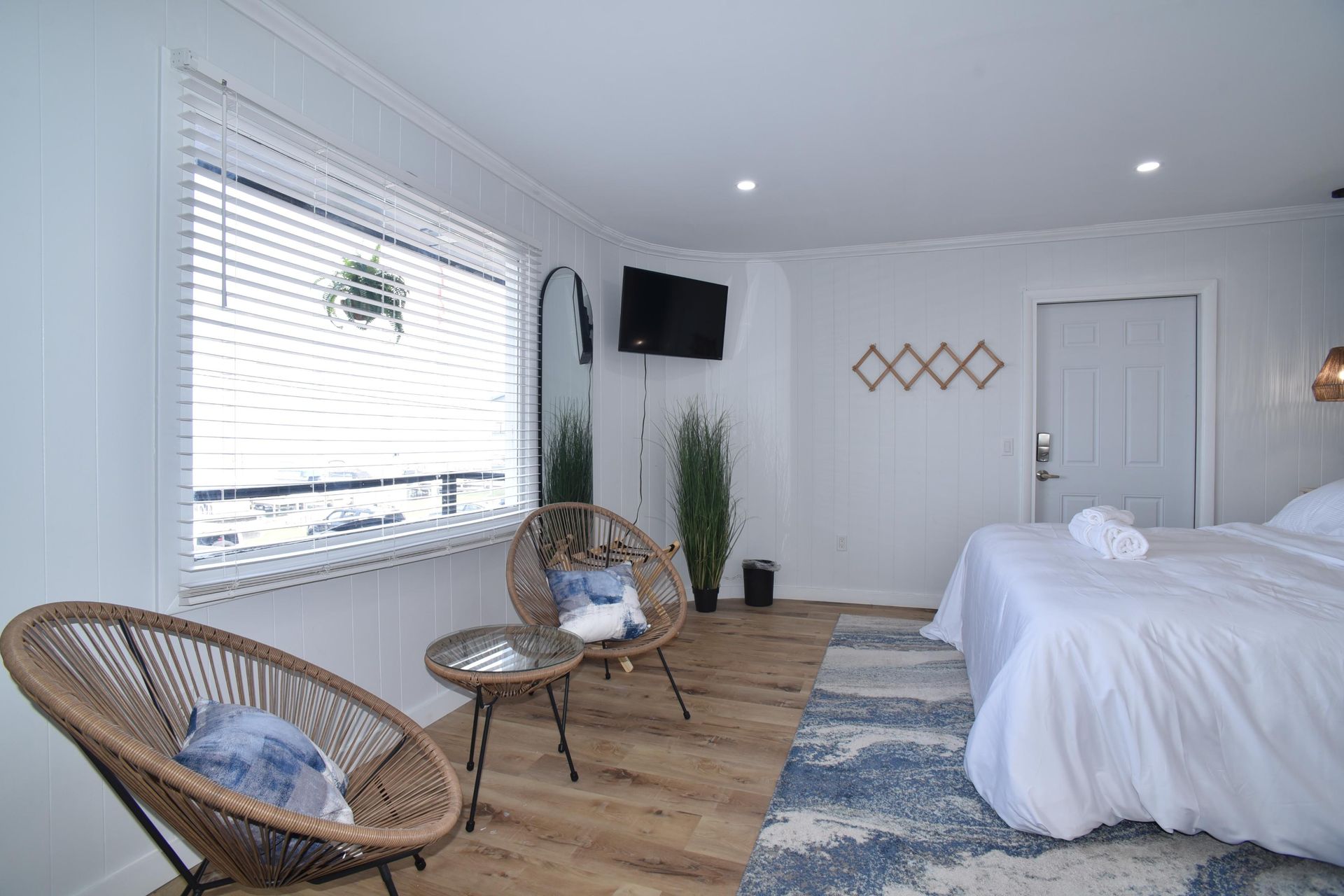 A bright hotel room with a white bed, two round rattan chairs, a small table, a wall-mounted TV, and a window with blinds.