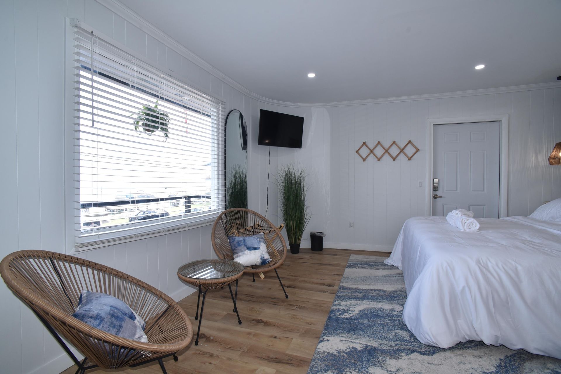 A white hotel room with two wicker chairs, a small table, a bed with white bedding, and a blue patterned area rug.