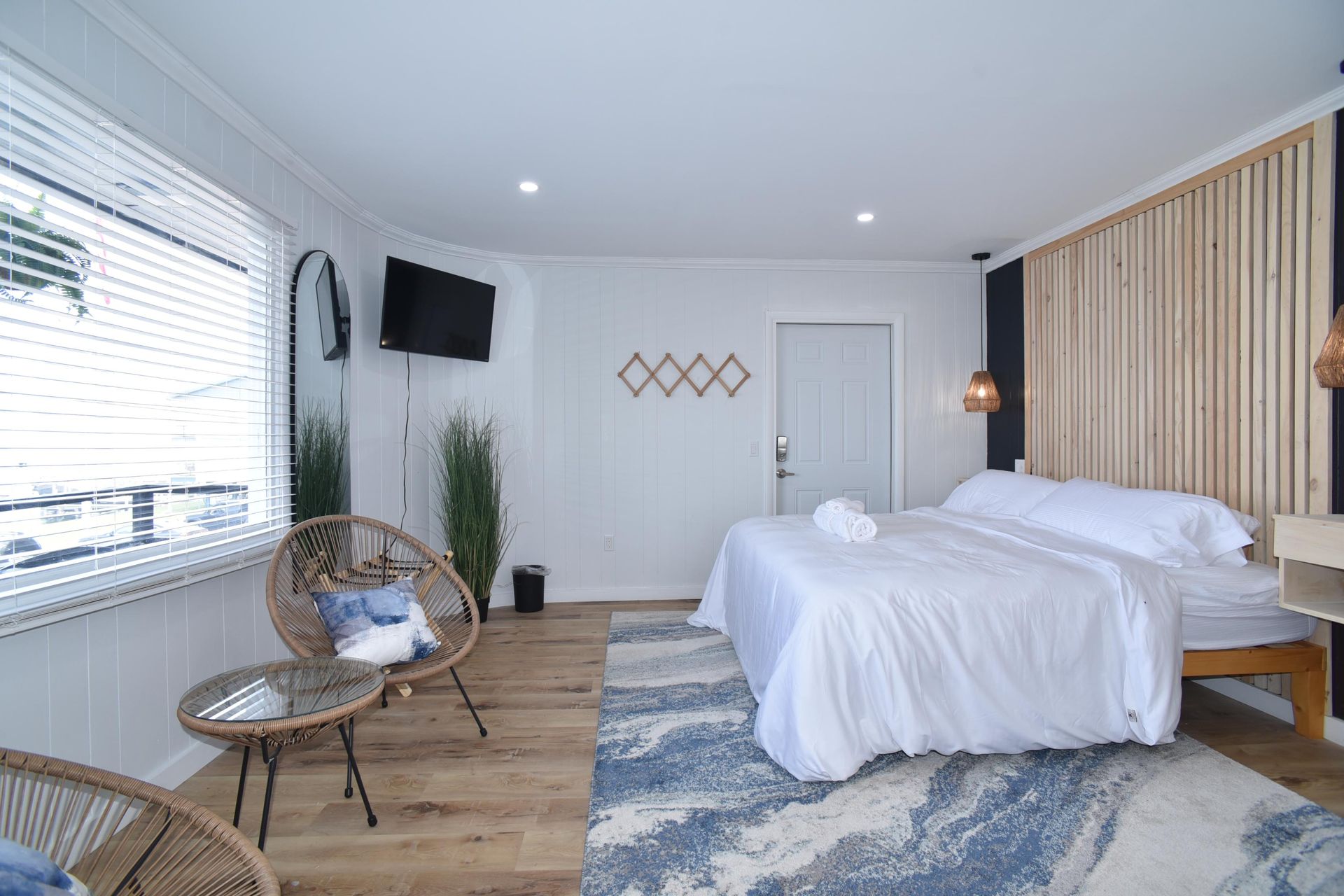 A bright bedroom with a white-made bed, a blue-and-white area rug, and two round wicker chairs by a window.