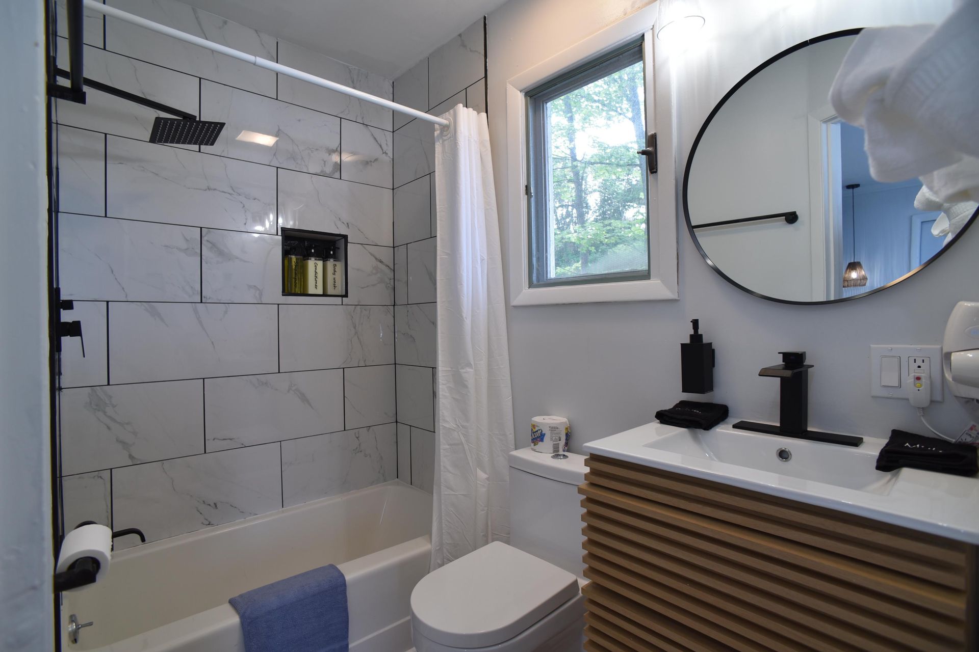 A modern bathroom featuring a white marble-tiled tub, a round mirror, and a slatted wood vanity with black fixtures.