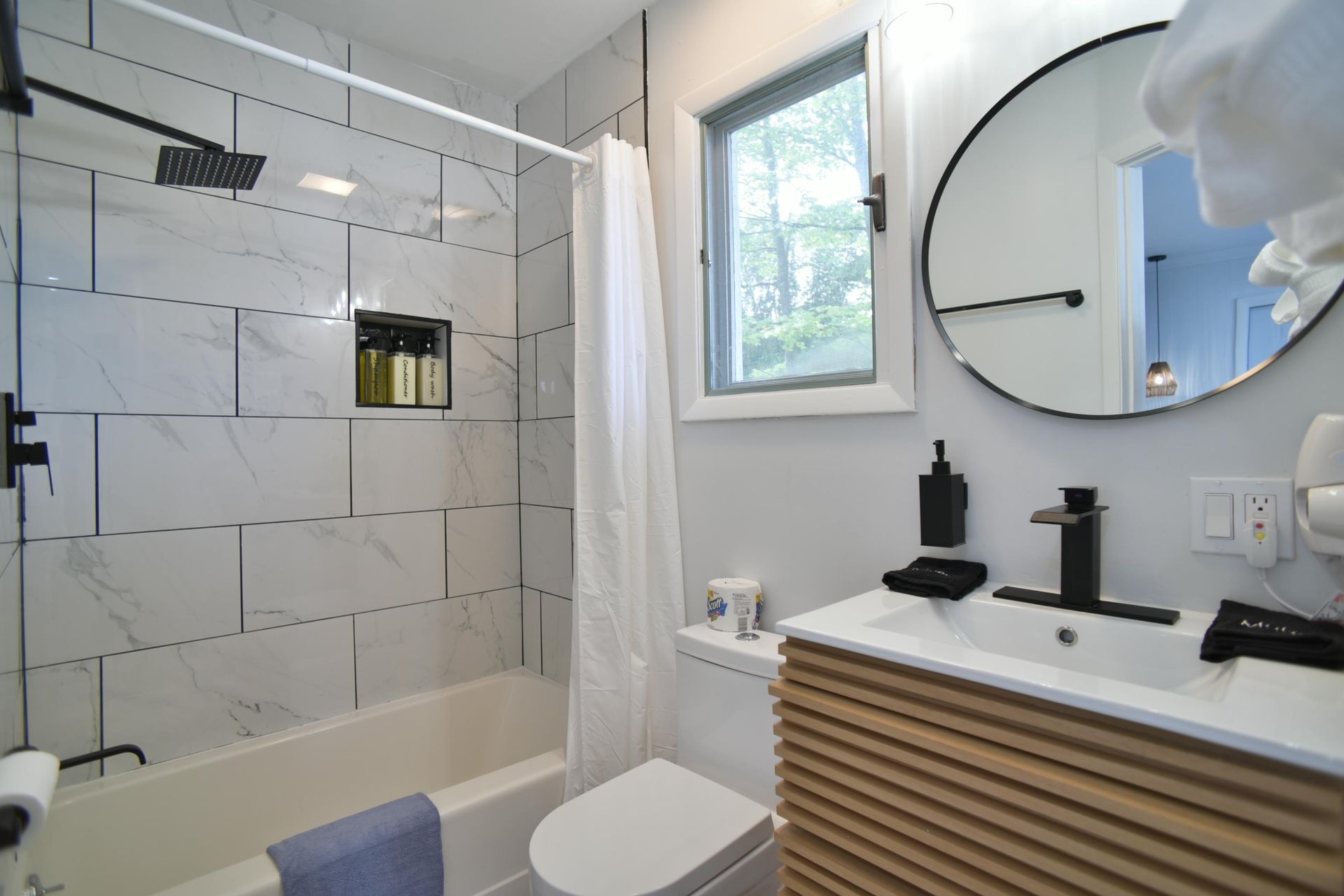 A modern bathroom with a white marble-tiled shower, wood-slatted vanity, round mirror, and a small window.