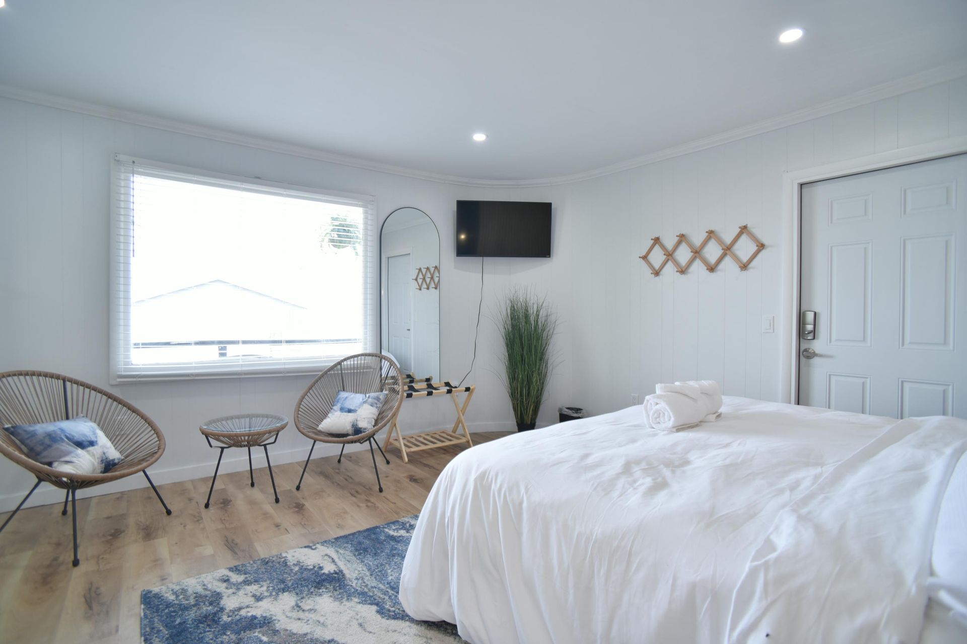 A bright, modern hotel room with a white bed, two wicker chairs, a small table, a wall-mounted TV, and a blue rug.