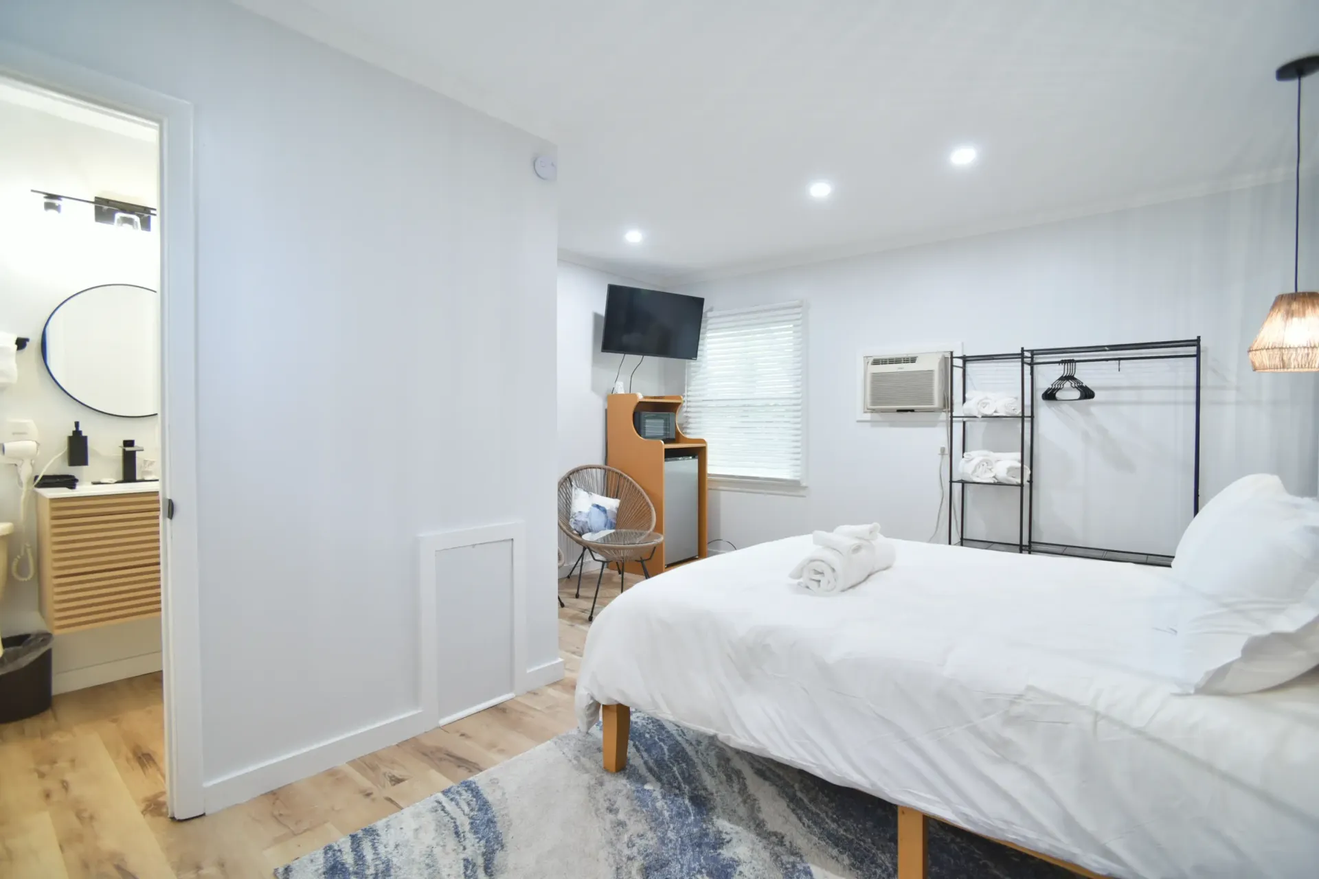 A modern, minimalist bedroom with white walls, light wood floors, a bed with white bedding, and a doorway to a bathroom.