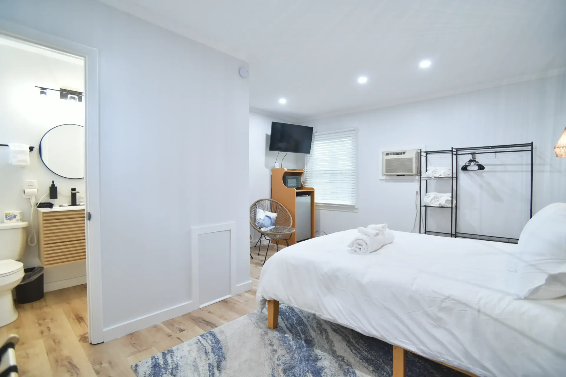 A bright, neutral-toned hotel room featuring a bed, an open clothing rack, a small desk area, and an attached bathroom.