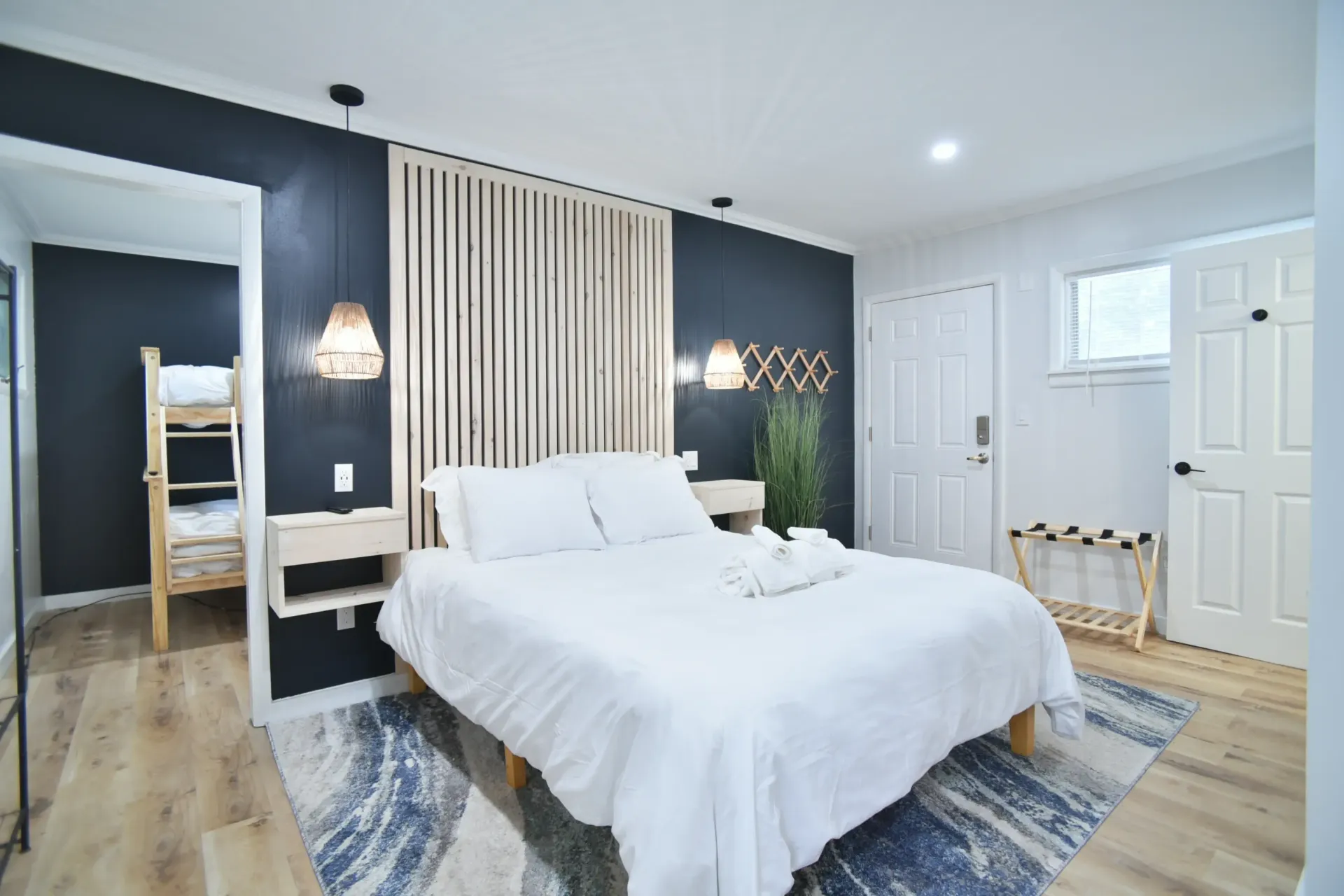 A hotel room featuring a queen bed with white linens, a wooden slat headboard, two pendant lights, and a bunk bed alcove.