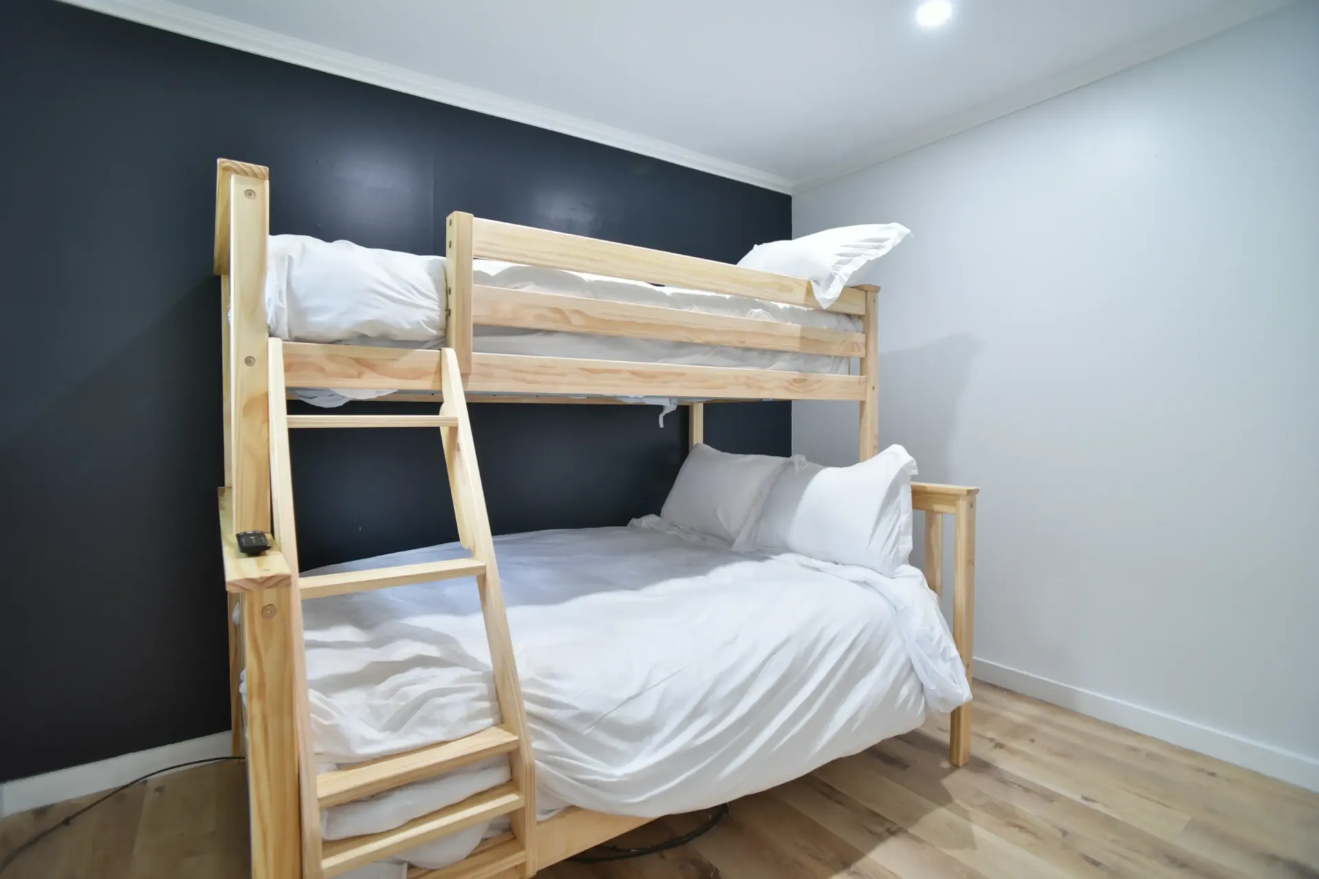 A light wood bunk bed with white bedding, positioned against a dark accent wall in a room with light flooring.