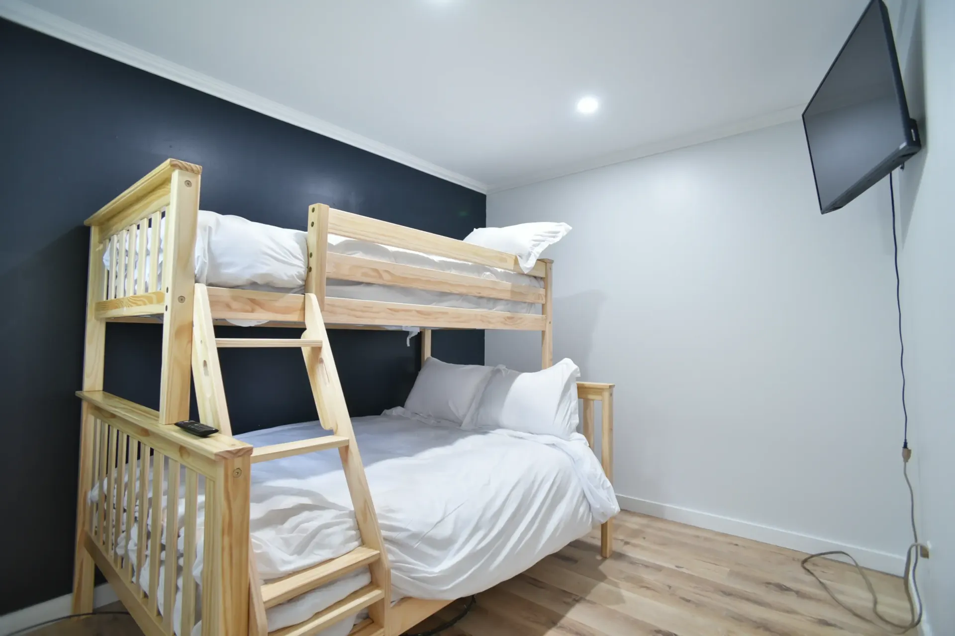 A light wood bunk bed with white bedding sits against a dark accent wall in a room with a wall-mounted television.