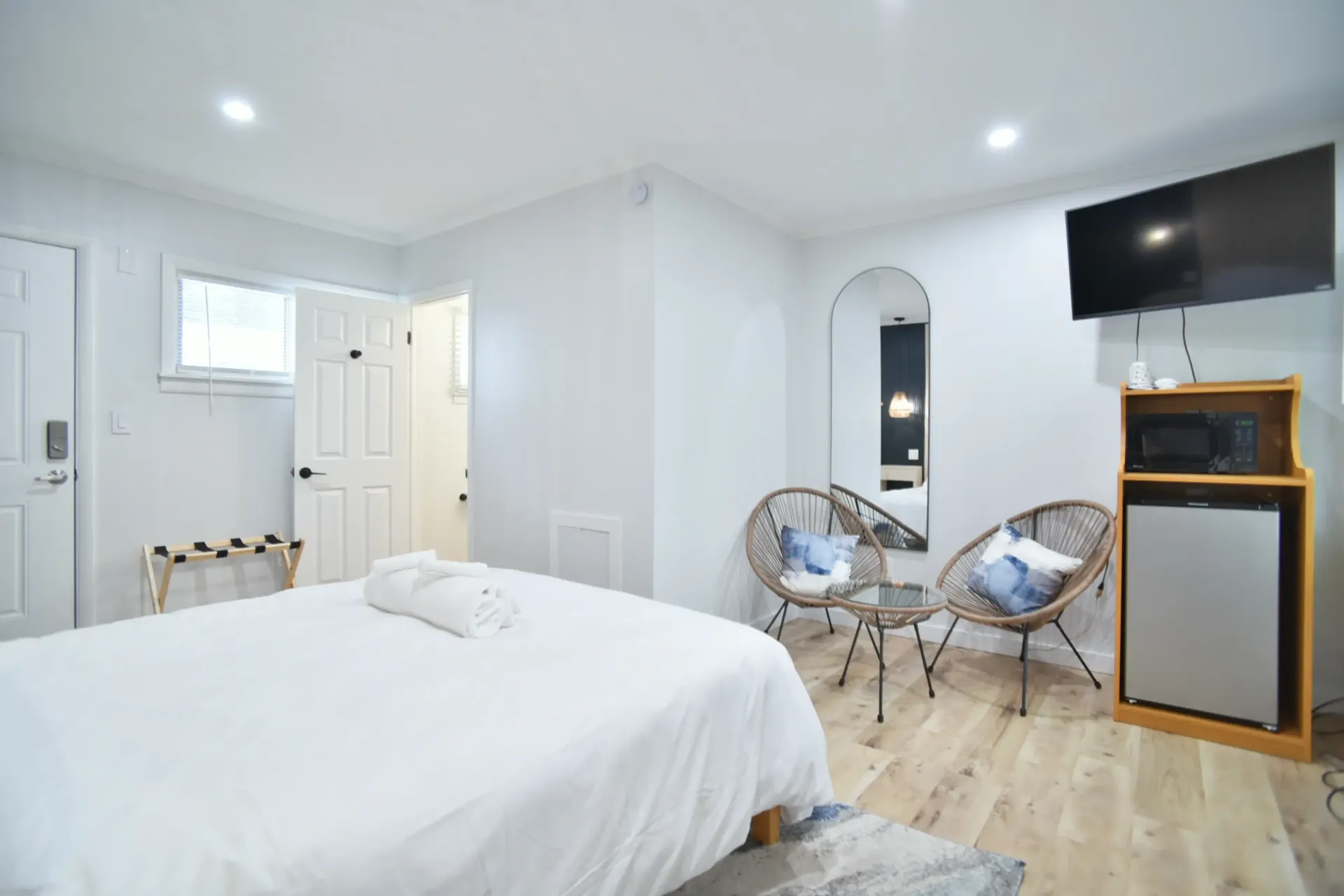 A modern hotel room with a bed, two round wicker chairs, a small table, a TV, and a mini-fridge on light wood floors.