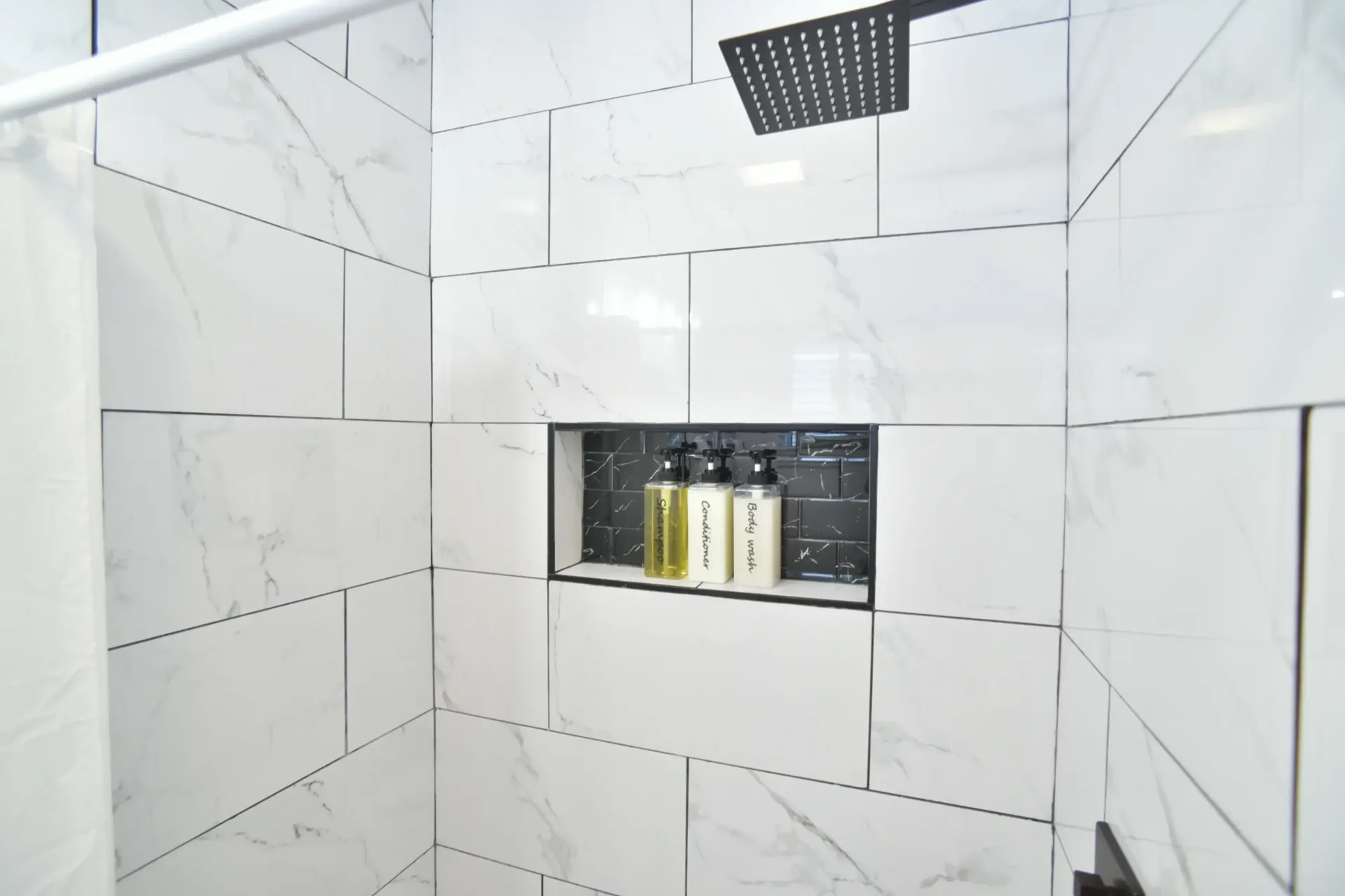 A shower stall with white marble-patterned tiles, a matte black shower head, and a recessed niche containing three bottles.