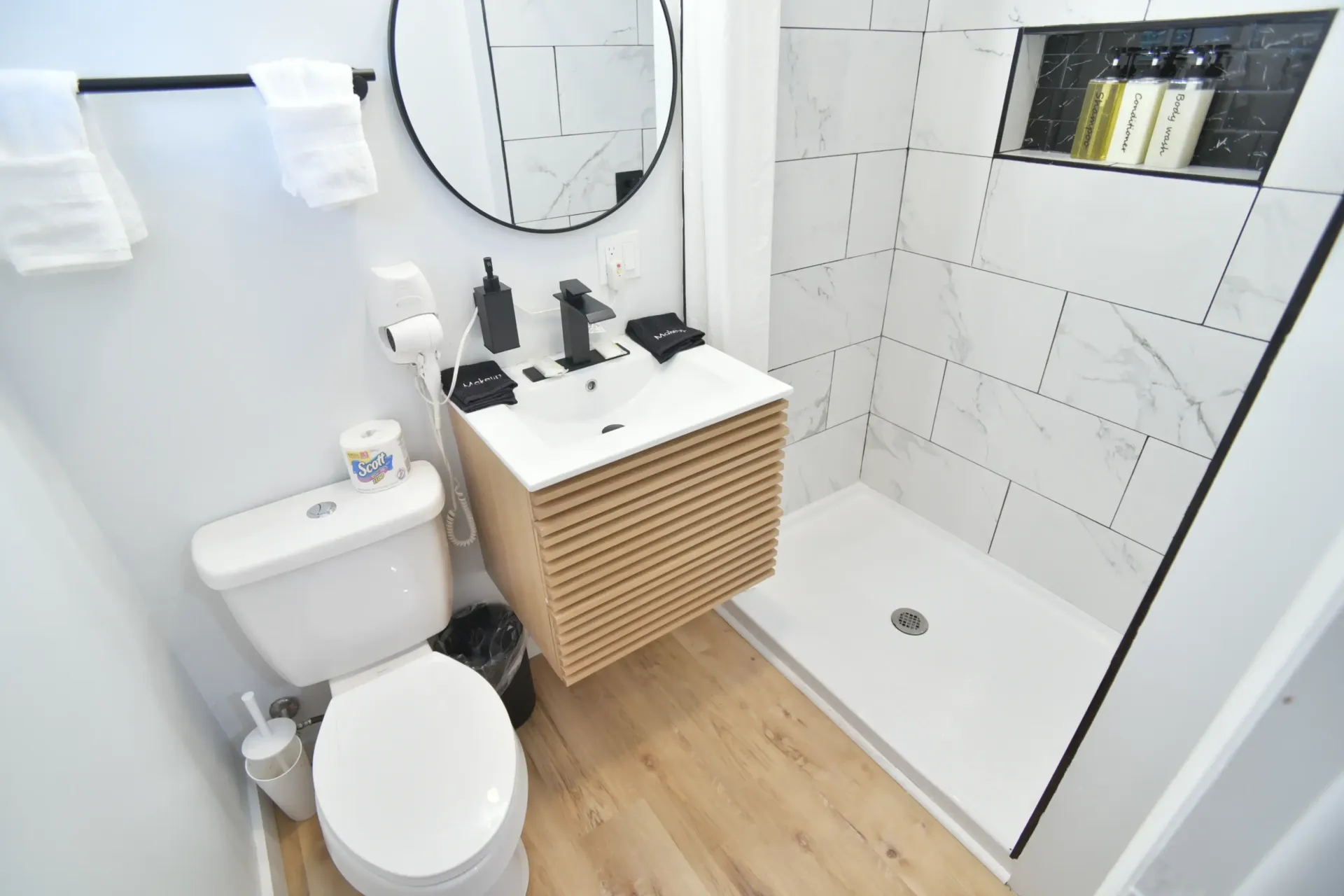 A modern bathroom featuring a white toilet, a wall-mounted wooden vanity with a sink, a round mirror, and a tiled shower.