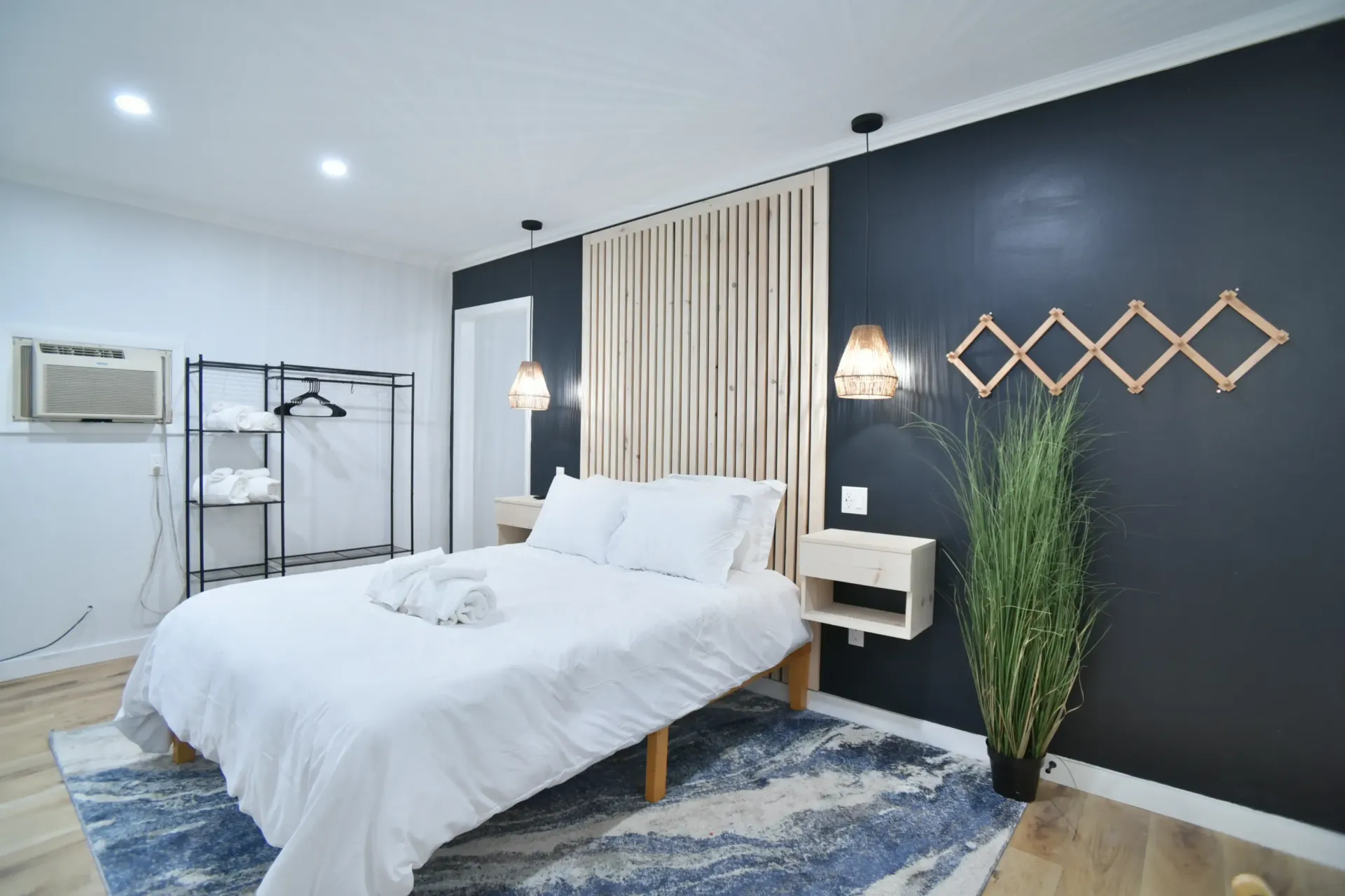 A minimalist bedroom featuring a white bed with blue rug, dark accent wall, wooden headboard, and two hanging lights.
