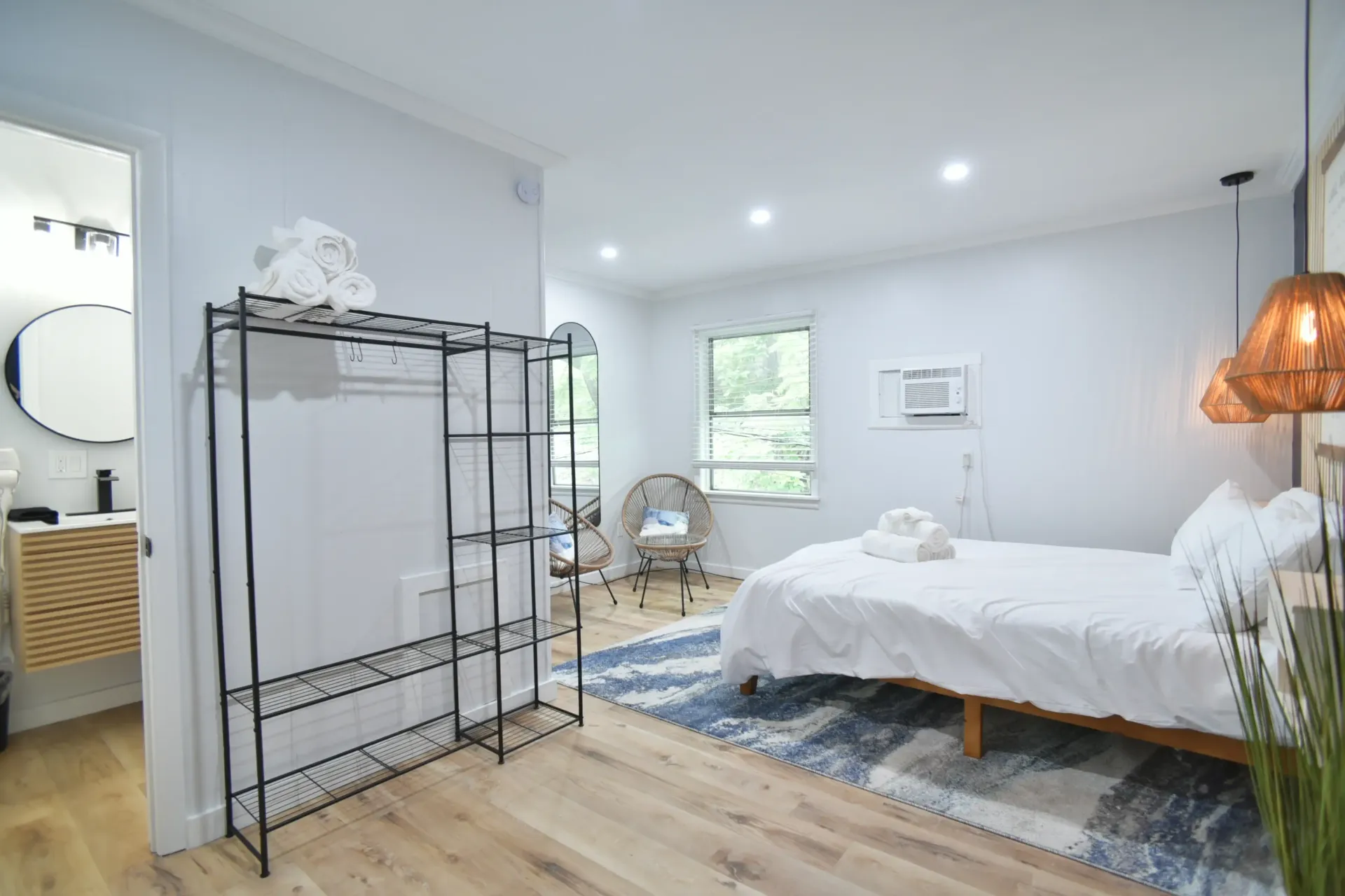 A bright, minimalist bedroom with a white bed, woven pendant lights, a metal clothing rack, and an adjacent bathroom.